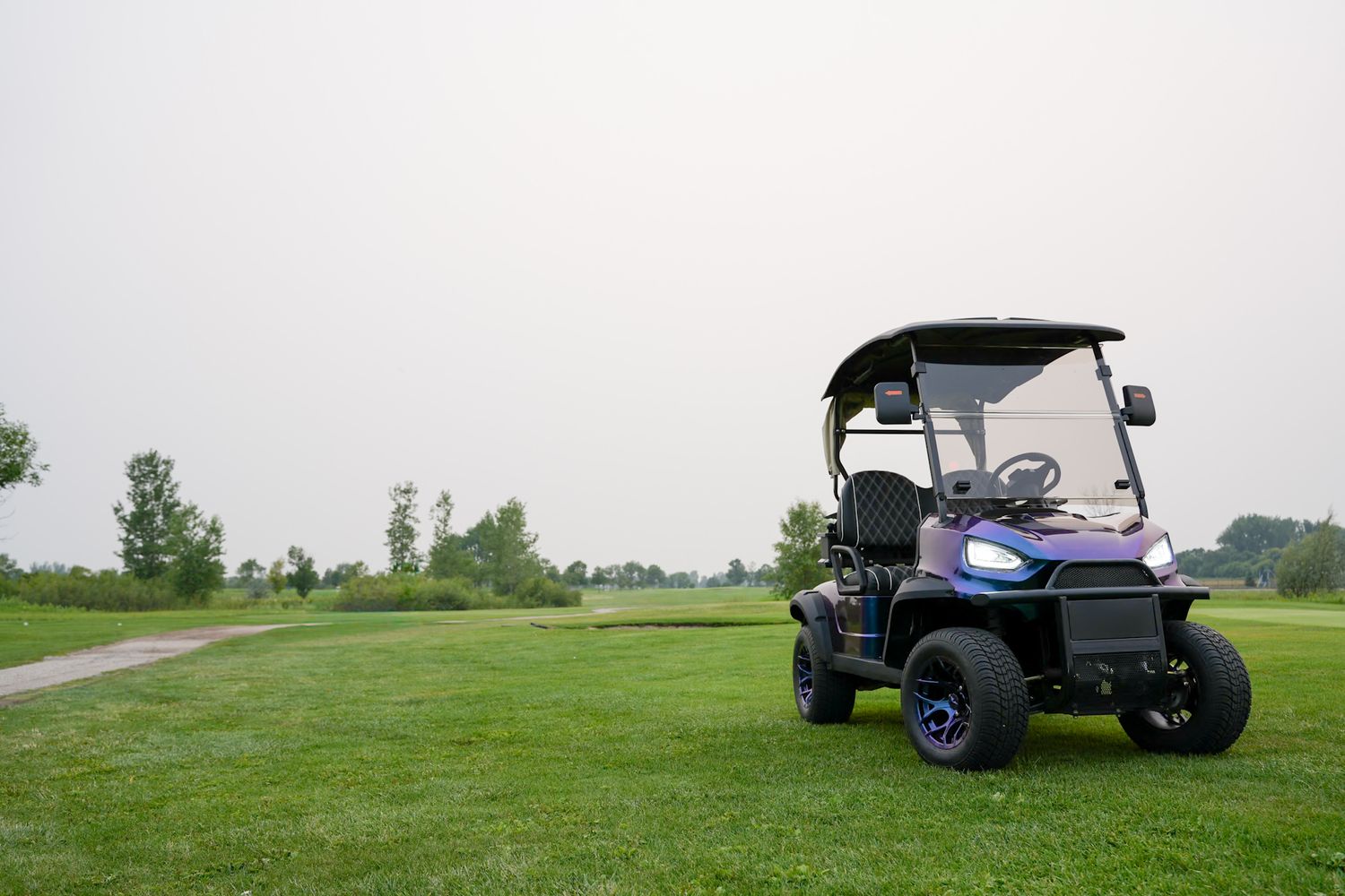 A golf cart driving on a lush green golf course with trees visible in the background on a sunny day.