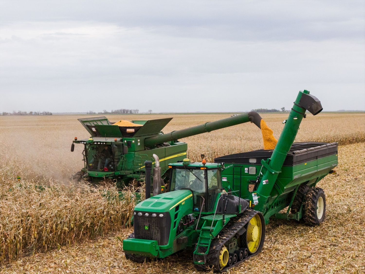 John Deere combine harvester and tractor with grain cart harvesting corn in a large farm field under cloudy skies.