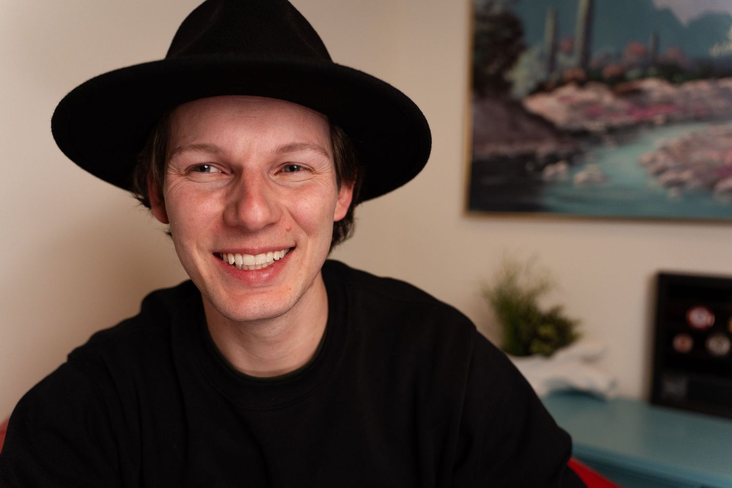 A person wearing a black hat smiles warmly while sitting indoors.