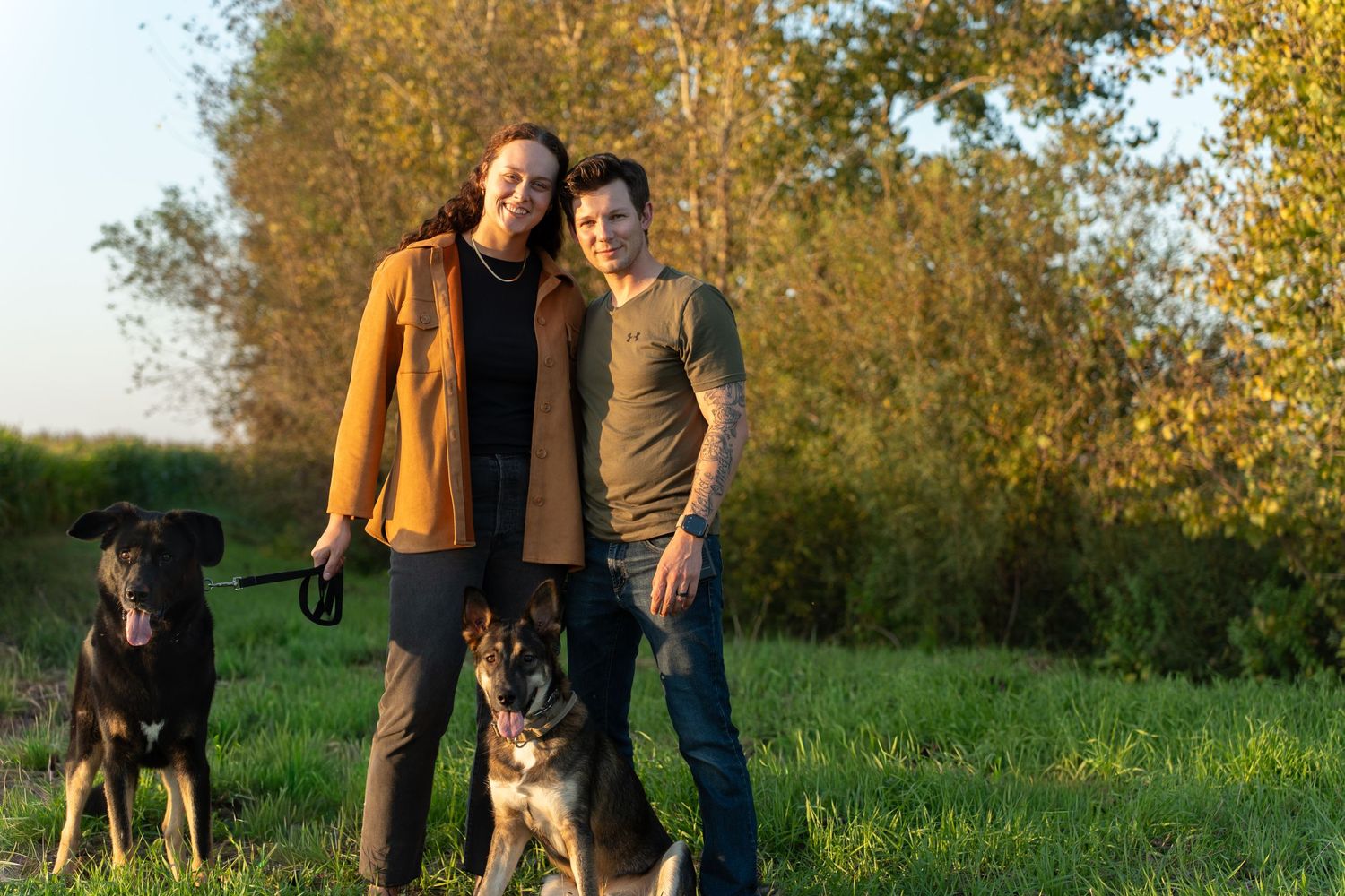 A couple walks their two dogs through a scenic autumn field during golden hour.