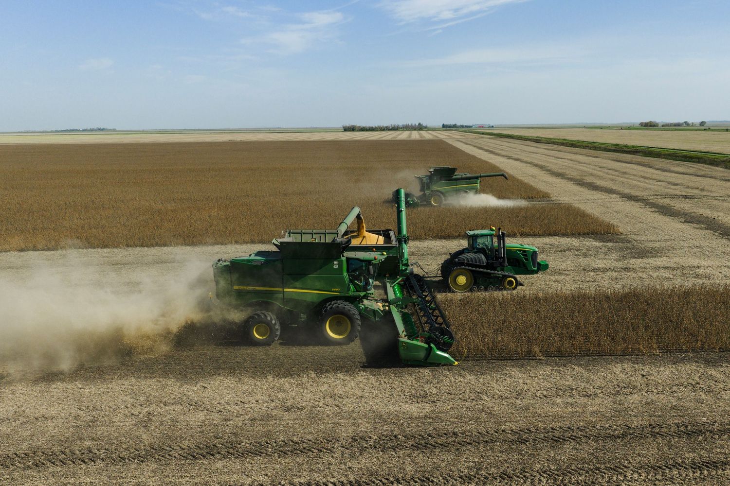 Agricultural machinery harvesting crops in a dusty field under blue skies.