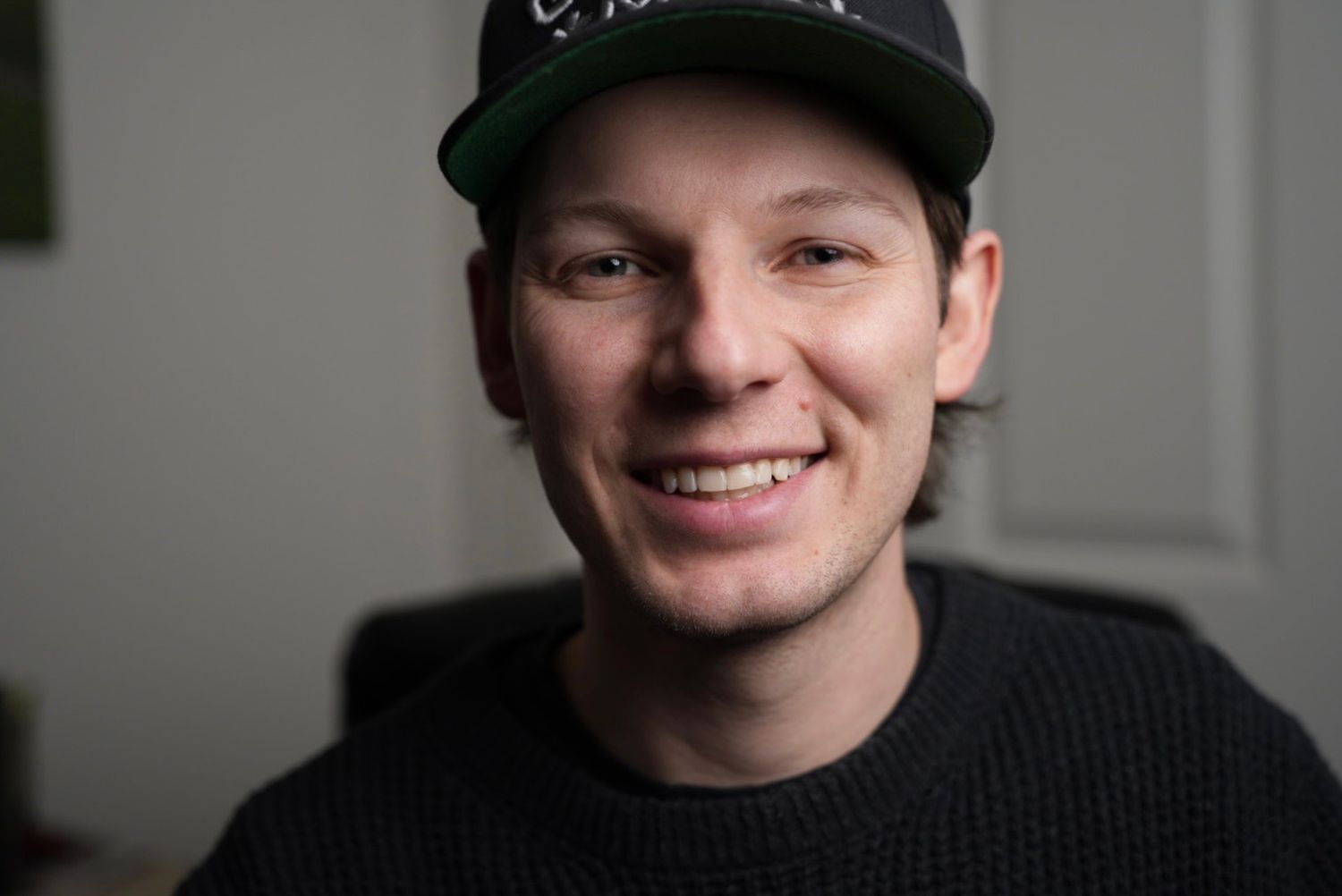 Someone wearing a black baseball cap and sweater smiles warmly at the camera in an indoor setting.