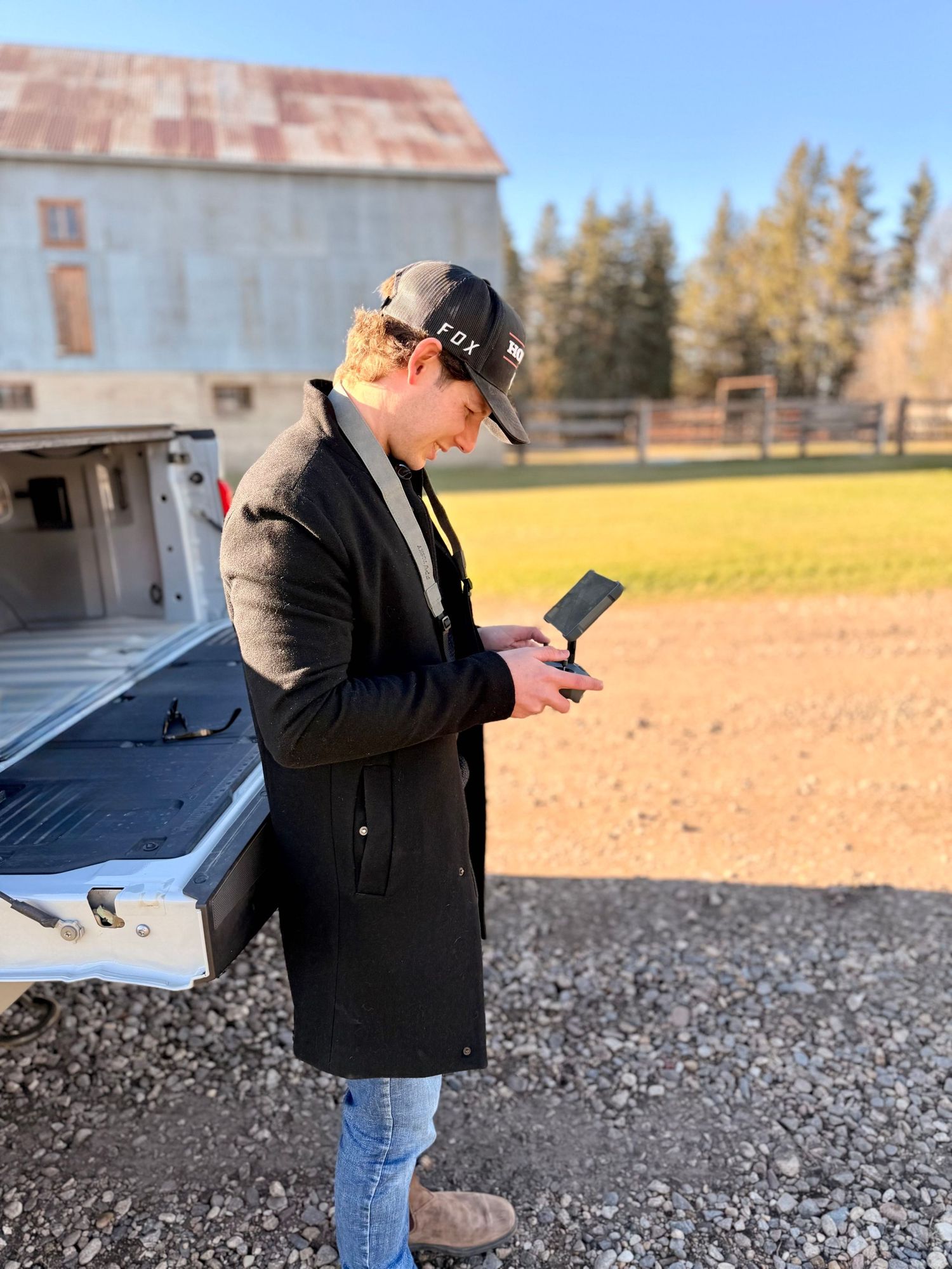 Person in black jacket and jeans taking measurements on a gravel surface with buildings in the background.