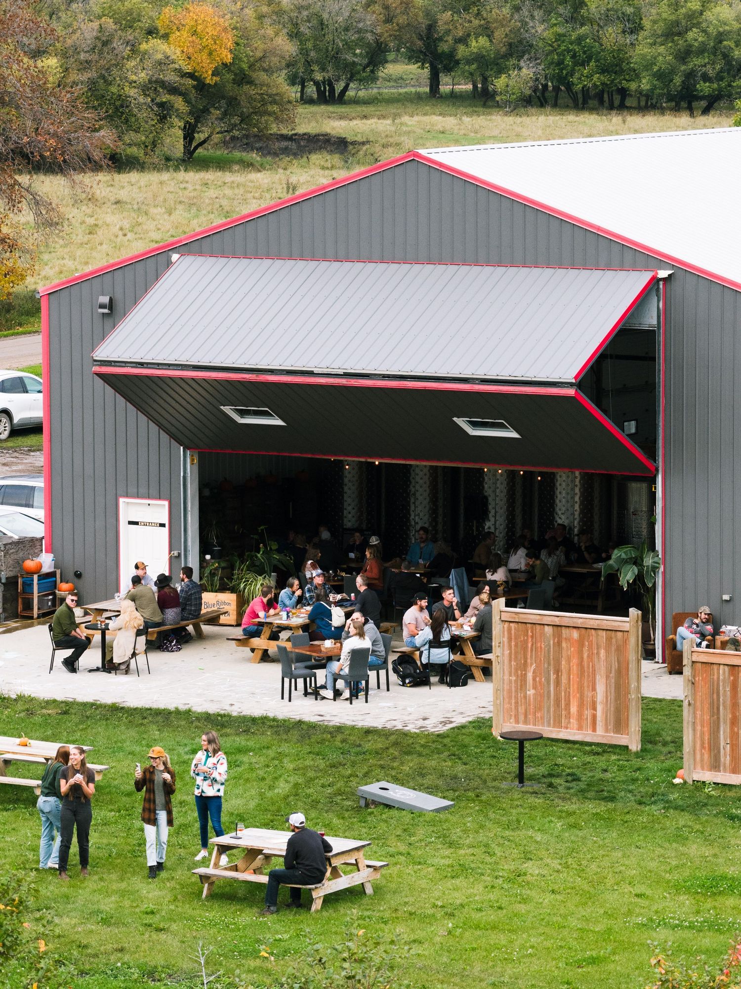 A modern pole barn venue with wooden accents and red trim hosts outdoor gatherings on a grassy lawn with picnic tables.