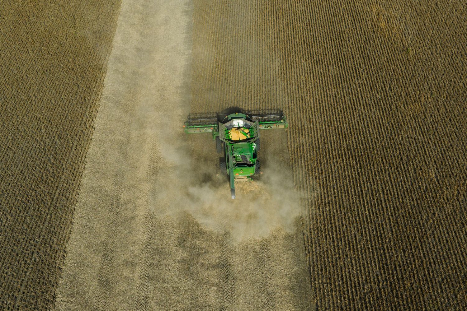 Aerial view of a green John Deere combine harvester working in a dusty agricultural field.