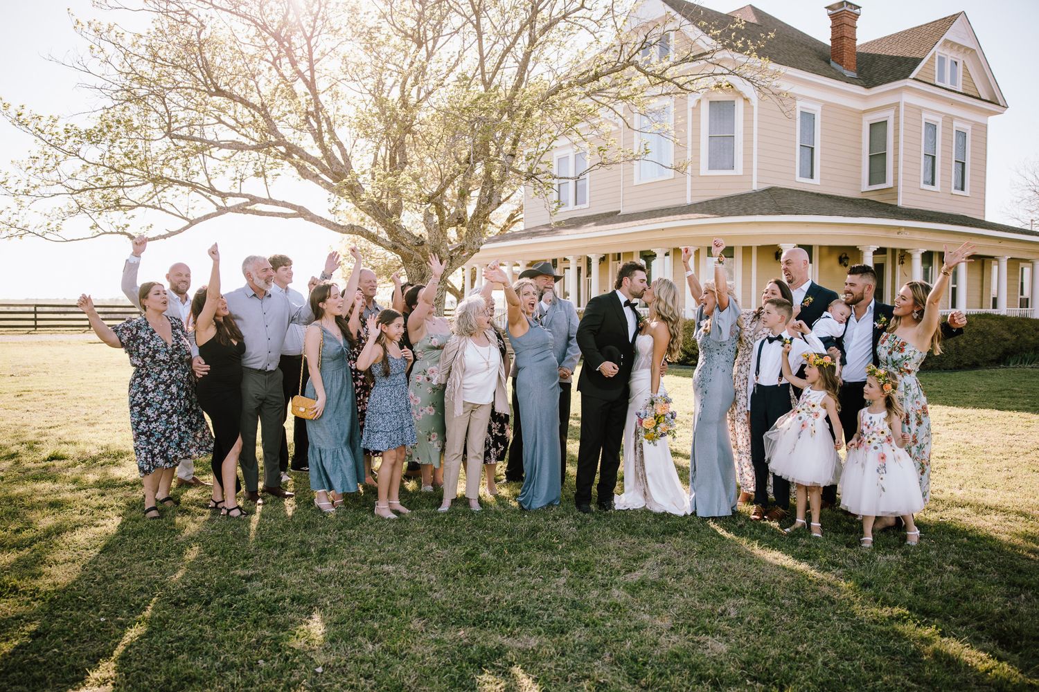 Wedding party celebrating outdoors in front of a historic farmhouse on a sunny day, bride and groom surrounded by family.