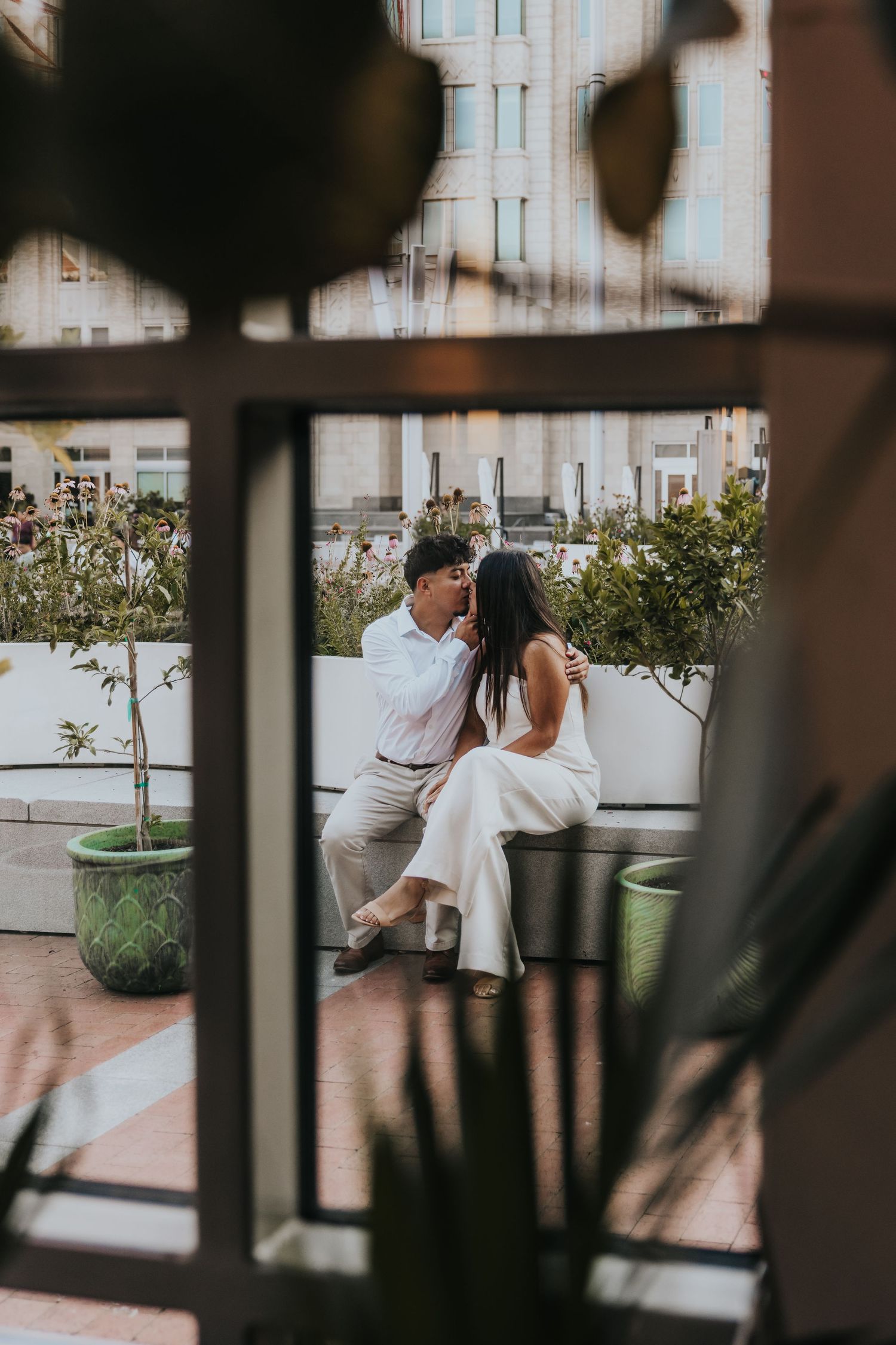 Romantic scene through window blinds with potted plants and outdoor seating visible.