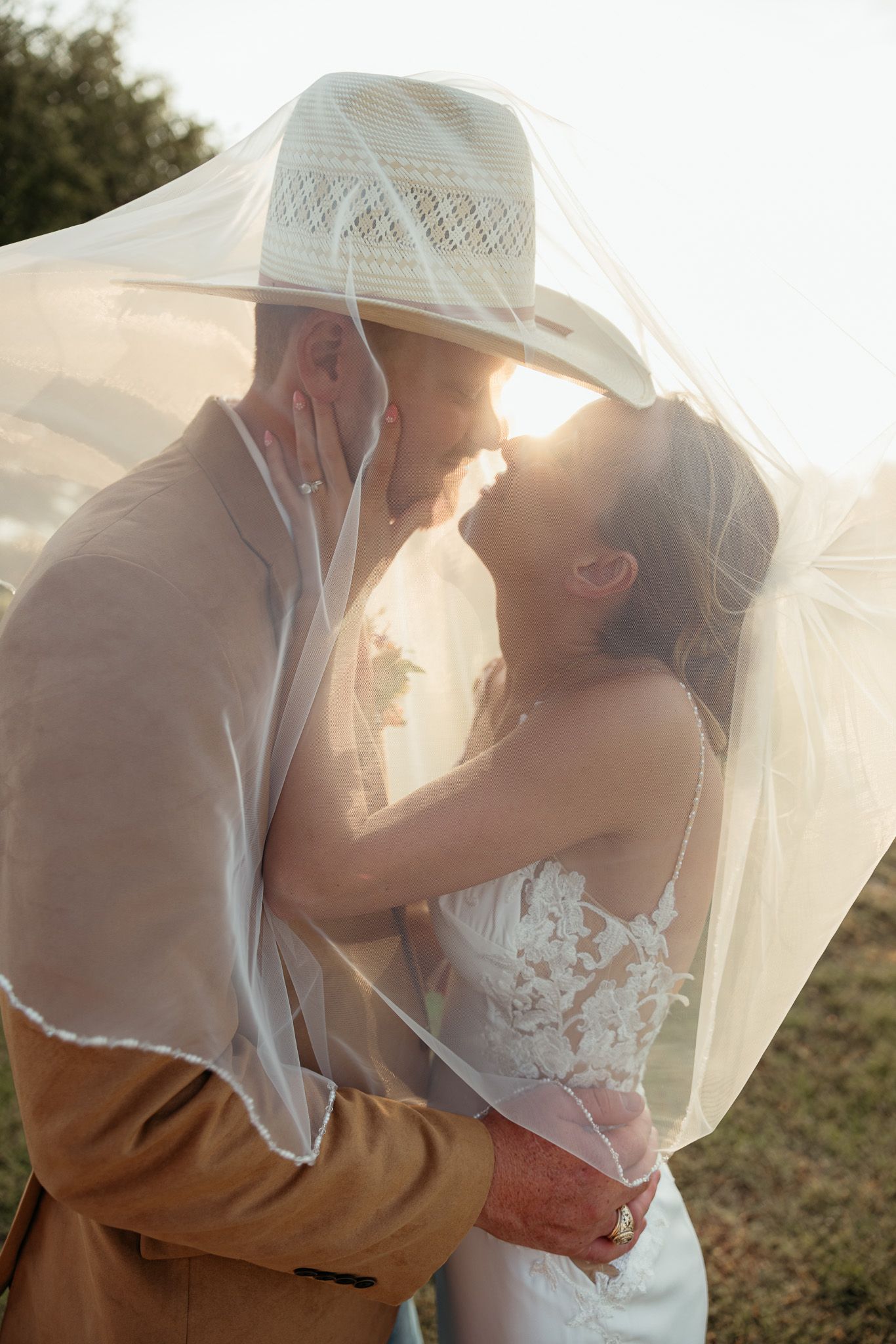 A romantic wedding couple embraces under a flowing veil during sunset creating a dreamy silhouette effect.