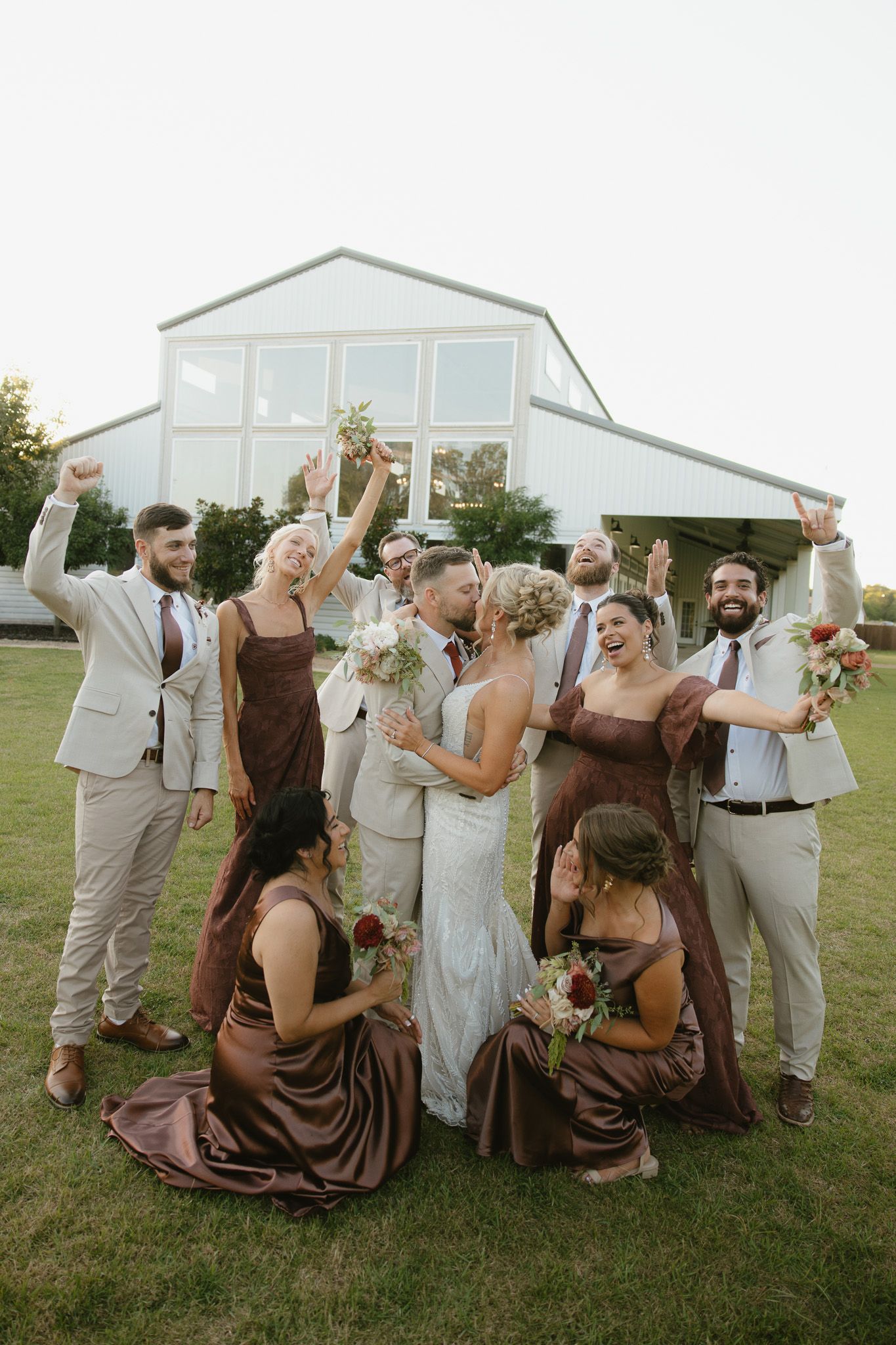 Wedding party celebrating outdoors with bride and groom kissing, bridesmaids in brown dresses and groomsmen in tan suits.