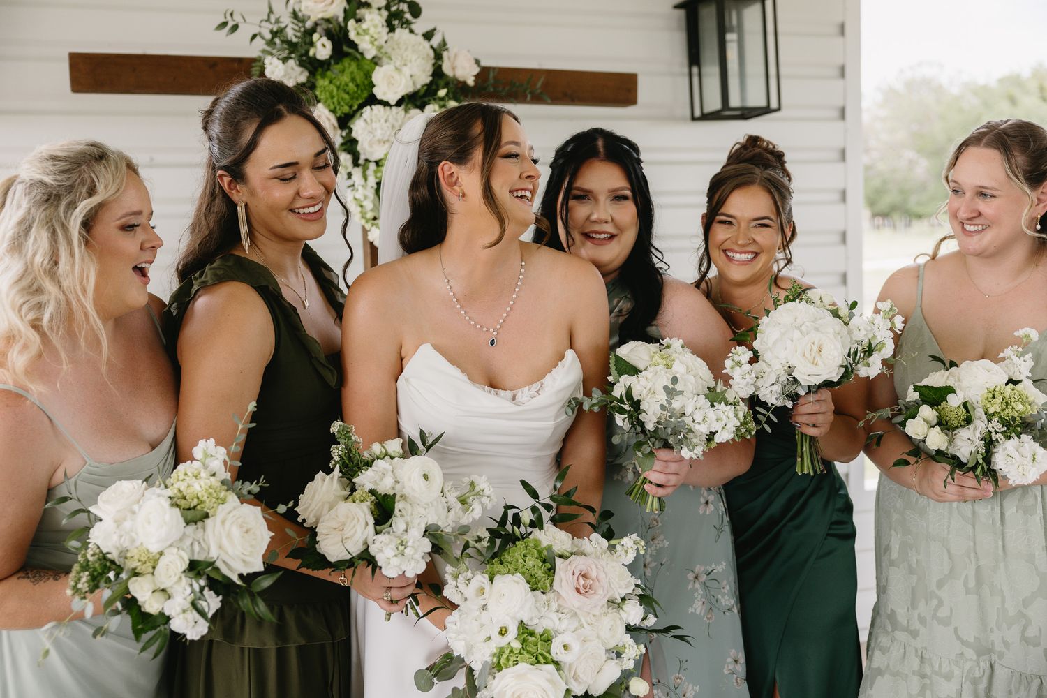 Bride in white gown laughing with bridesmaids in green dresses holding white floral bouquets outdoors.