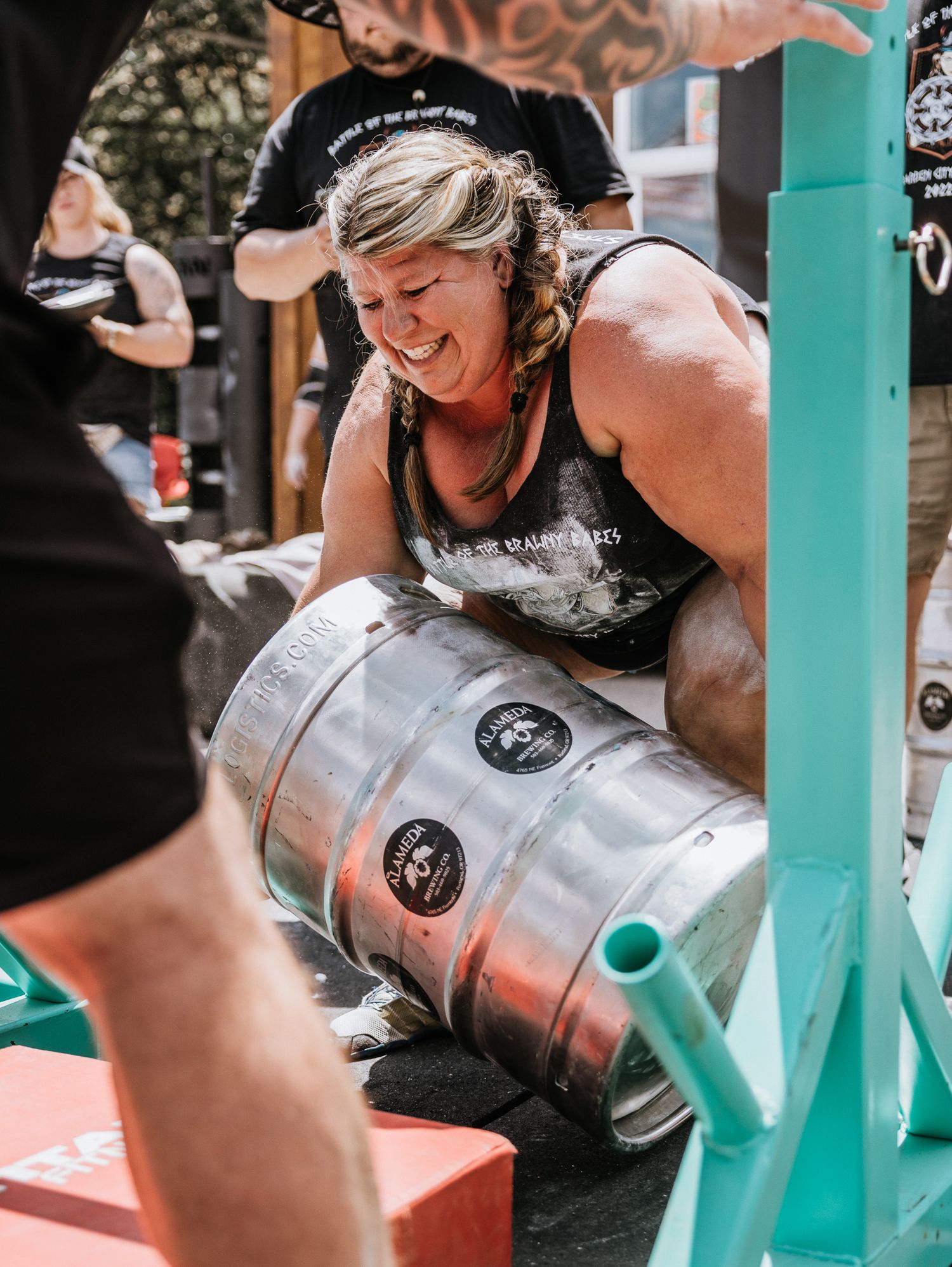 Athletes competing in a keg lifting competition drinking from metal kegs at an outdoor event.