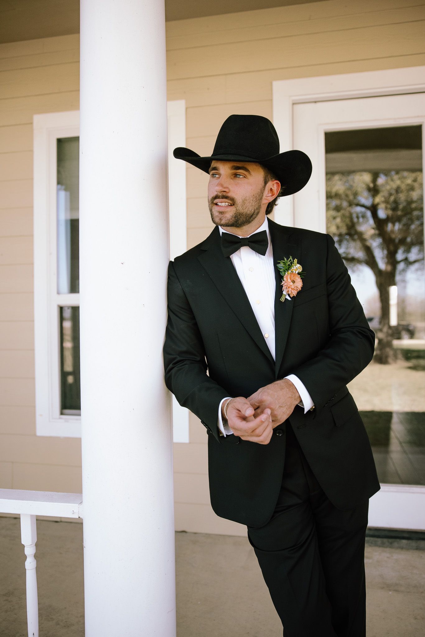A groom in a black tuxedo and cowboy hat with a boutonniere leans against a white porch column.