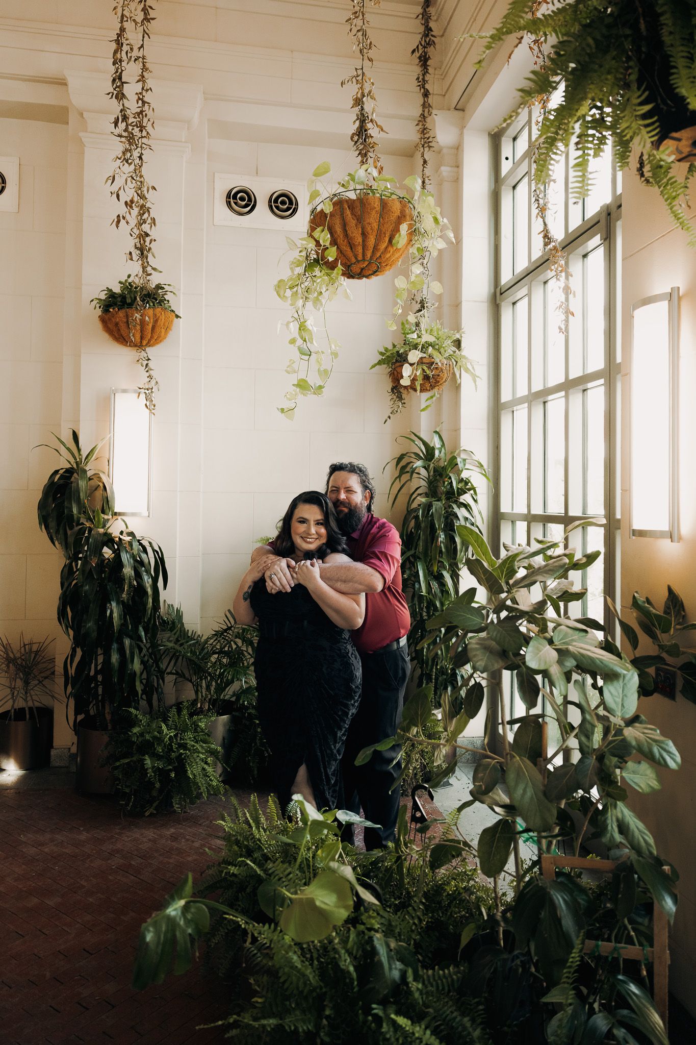 Couple embracing and smiling among lush indoor plants with hanging baskets in a bright room with large windows.