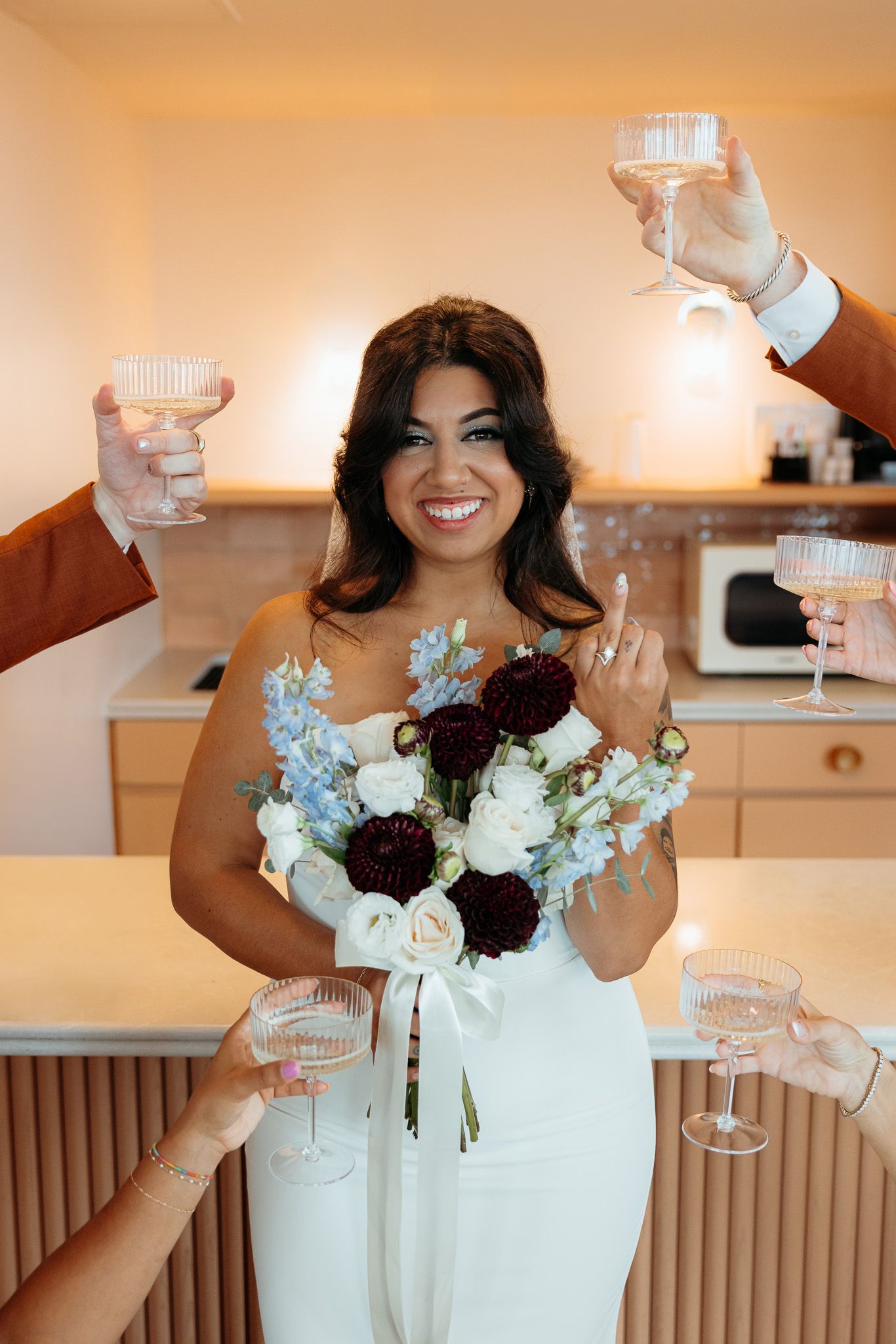 Smiling bride in white dress holds bouquet of white, blue, and burgundy flowers while guests raise champagne glasses in toast.