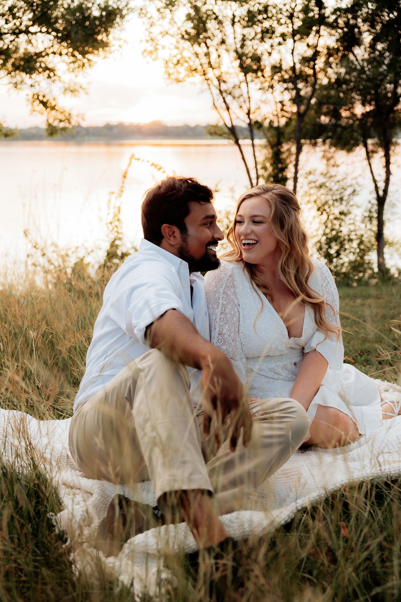 A romantic couple in white clothing laughs together while sitting in tall grass at sunset near a lake.