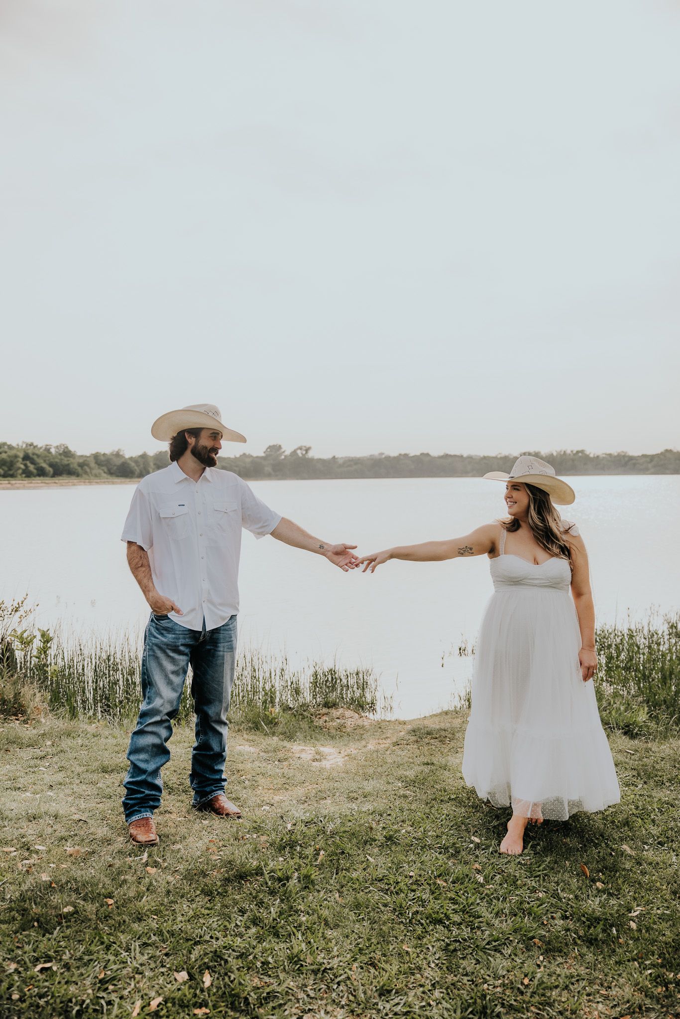 A couple in wedding attire holds hands while standing in a grassy area near a serene lake on their special day.