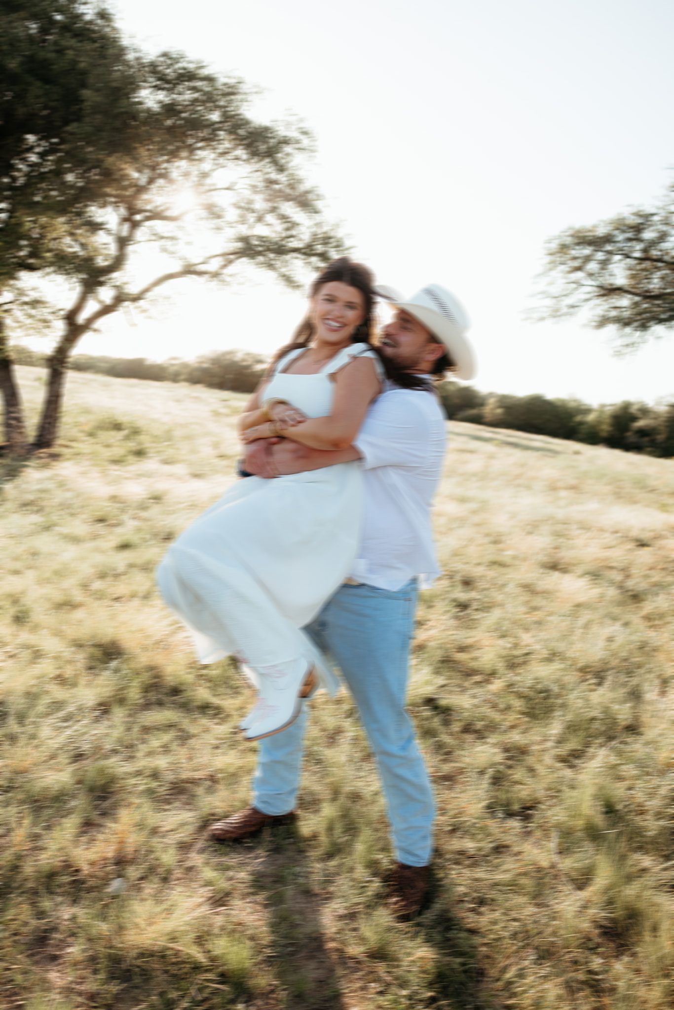 A joyful couple in white attire embraces in a sunlit field, the man lifting the woman as they laugh together.