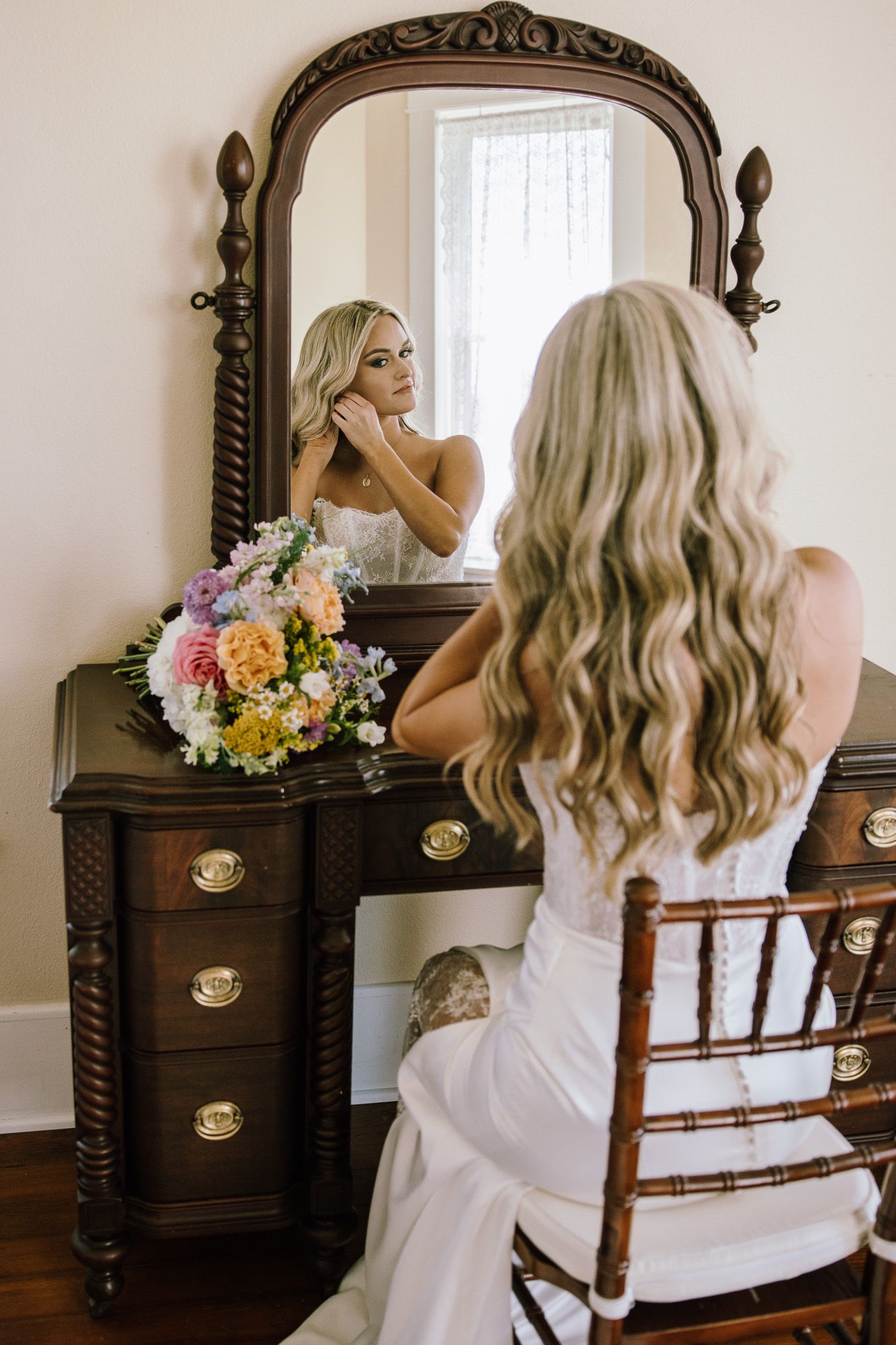 Blonde bride in wedding dress sits at vintage vanity mirror adjusting earrings with colorful floral bouquet nearby.