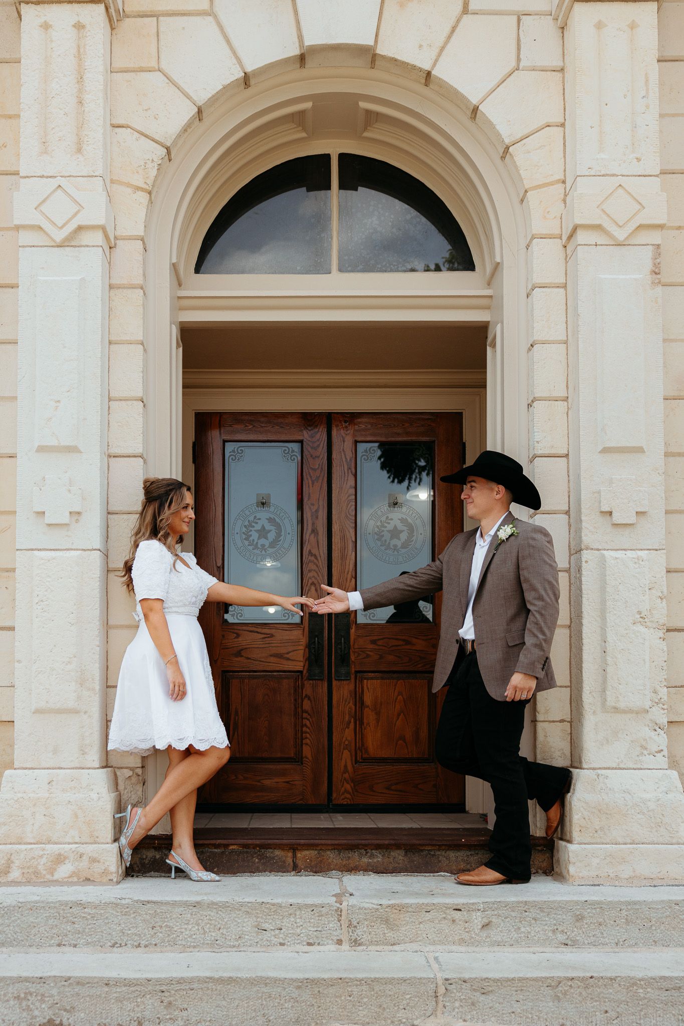 Couple in wedding attire holding hands at ornate wooden doors, bride in white dress, groom in cowboy hat and suit.