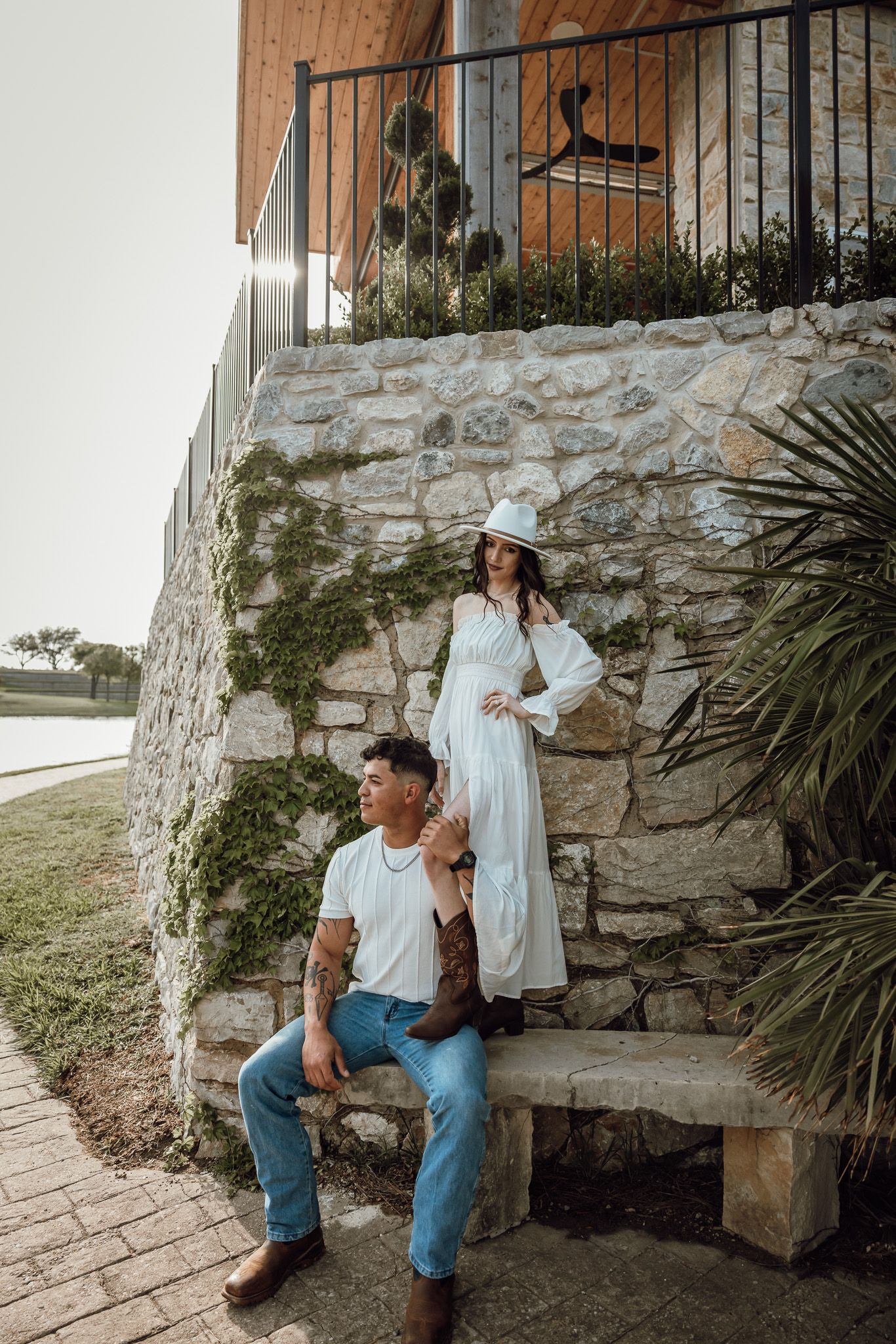 Couple sitting against a rustic stone wall with wooden railing and plants.