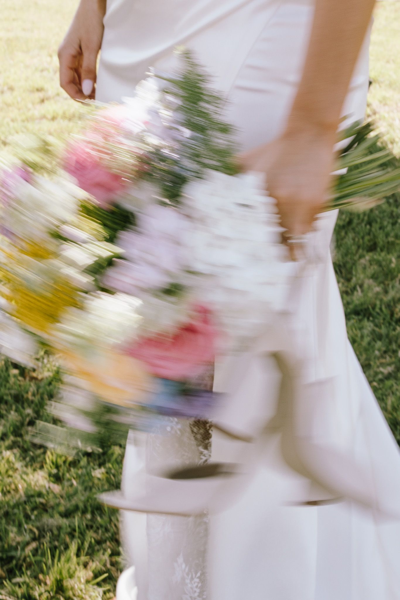 Bride in white gown holding colorful wedding bouquet with pink, yellow, and white flowers, captured in motion blur outdoors.