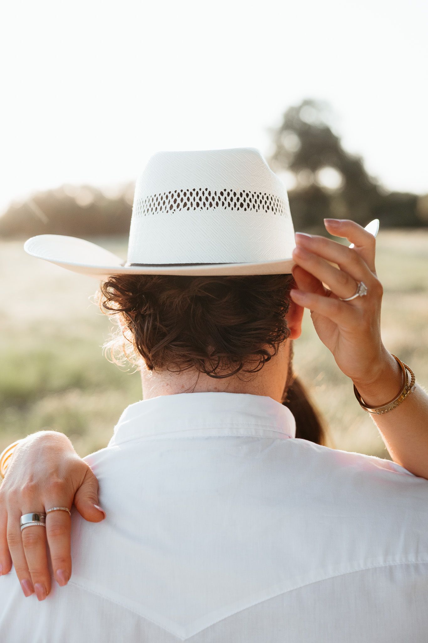 Couple embracing outdoors at sunset, woman touching man's white cowboy hat, both wearing wedding rings in a golden field.