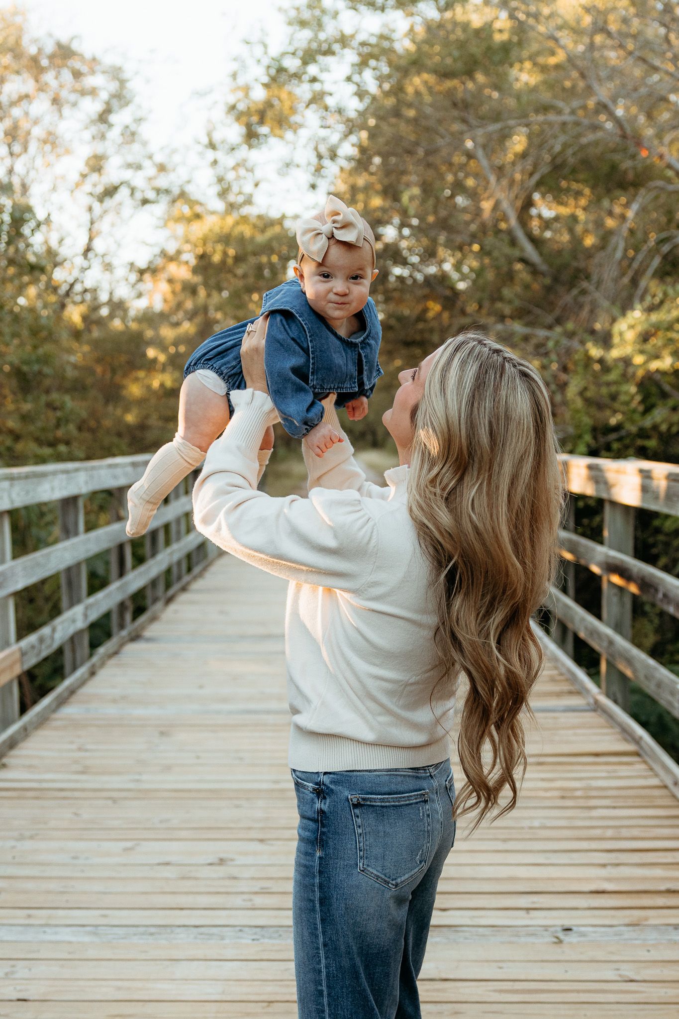 Mother lifting young child playfully on wooden boardwalk during sunset photo session.
