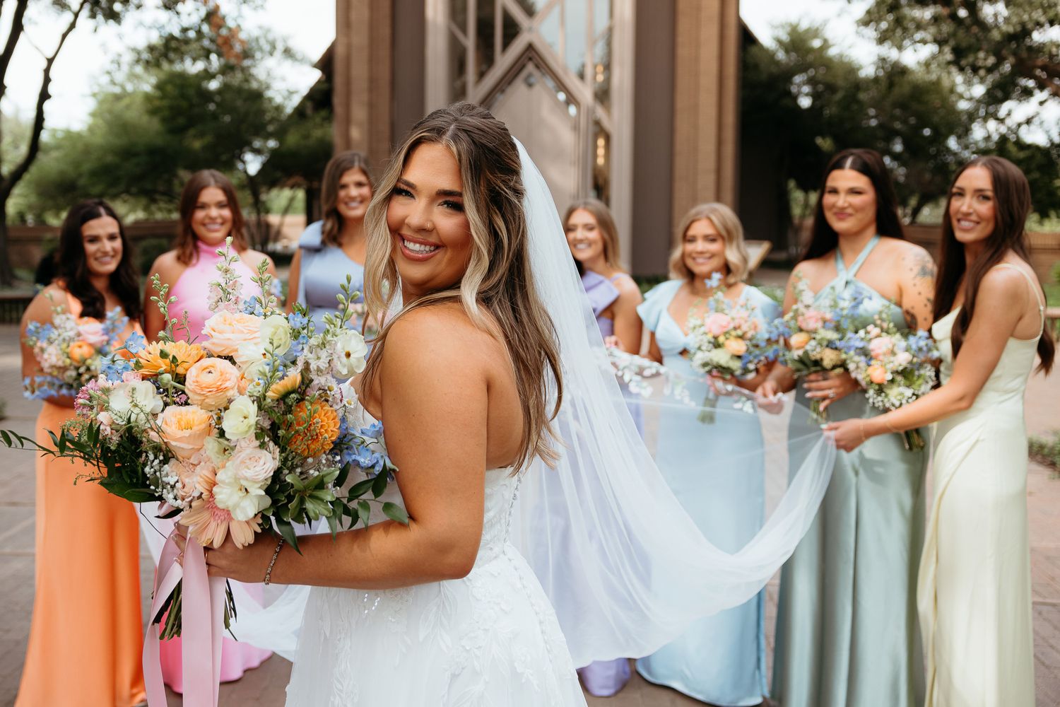 Bridesmaids in light blue dresses gather together holding colorful bouquets during an outdoor wedding celebration.