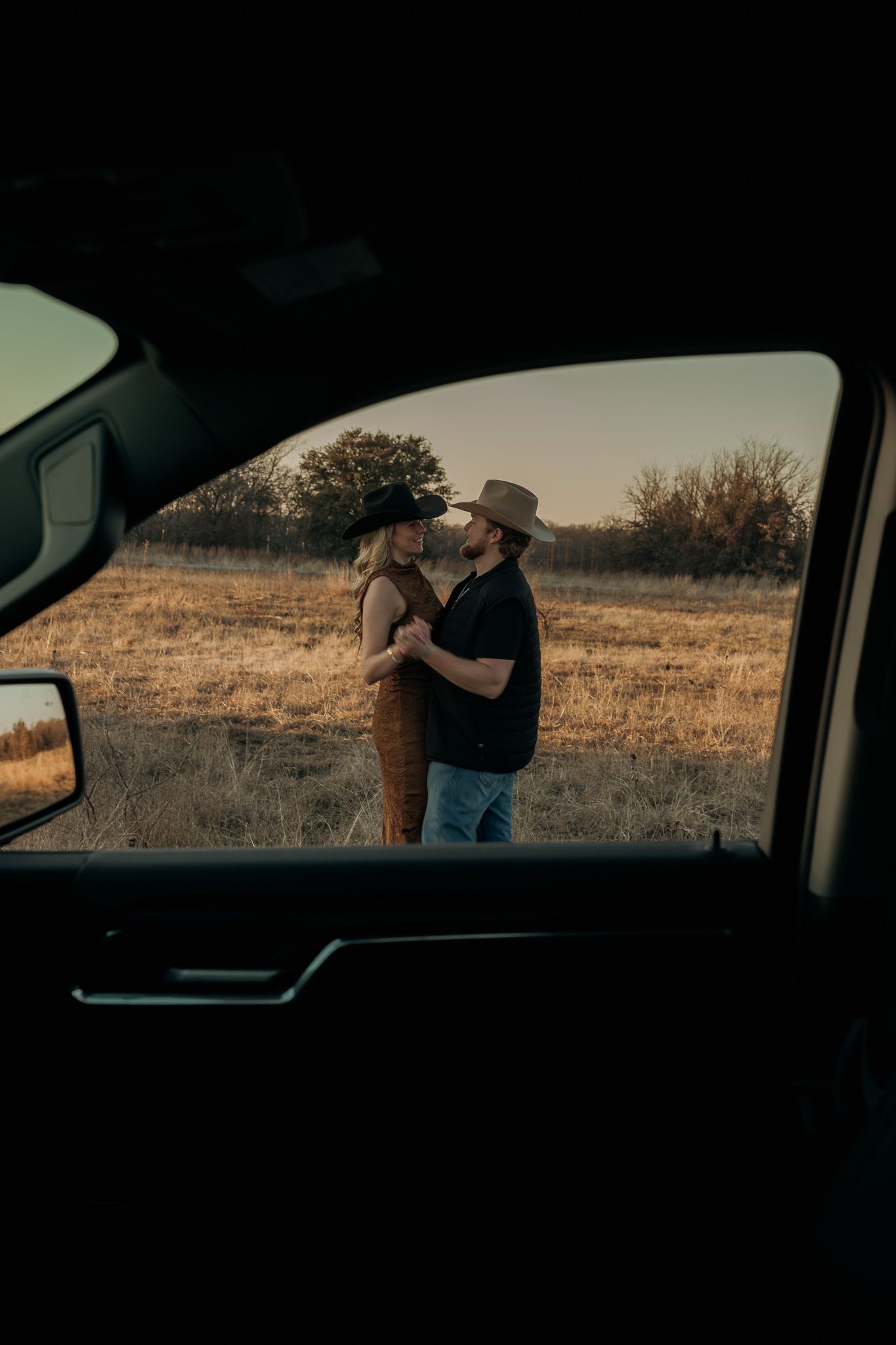 A romantic sunset moment viewed through a car window, with two figures embracing in a rural field at golden hour.