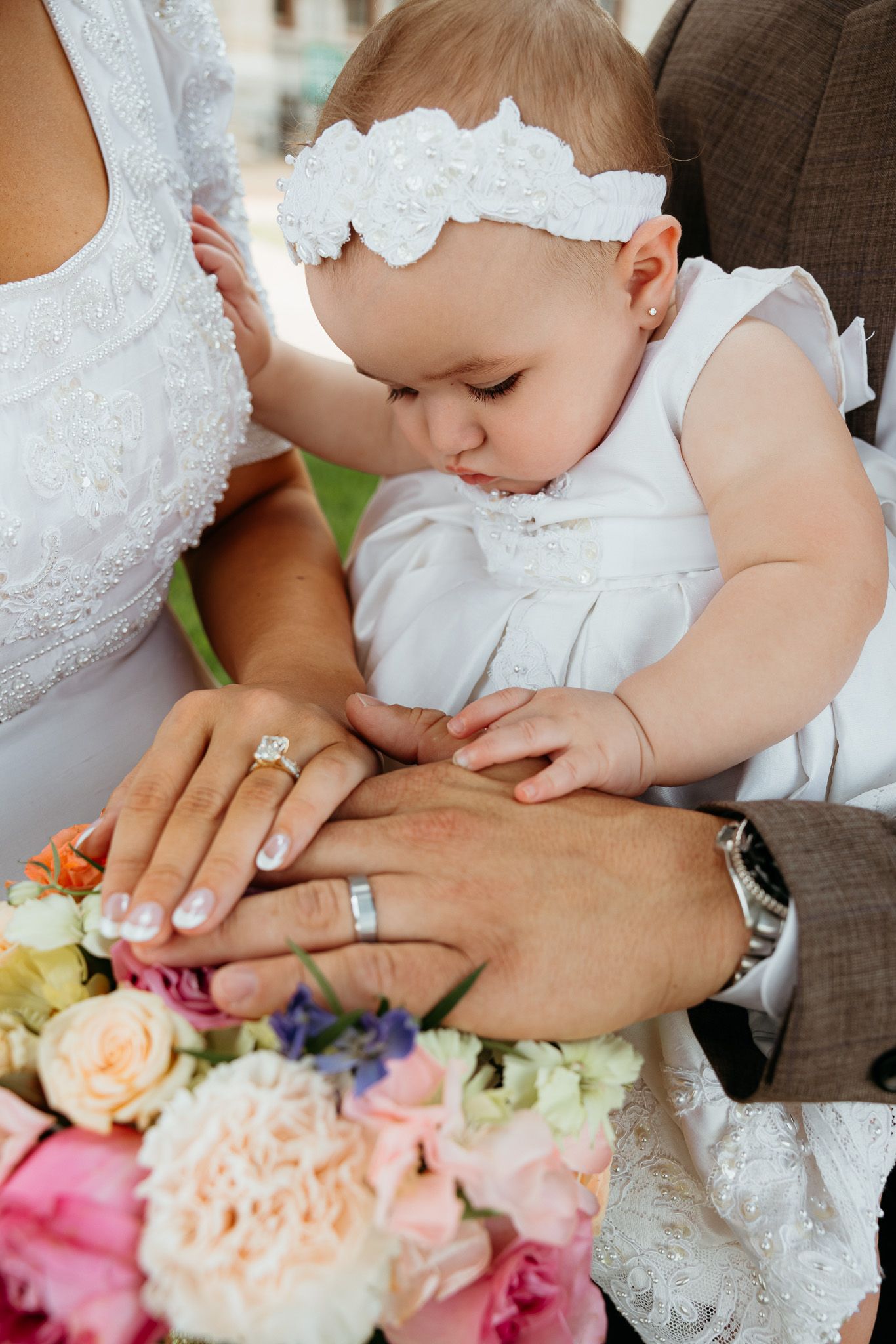 Baby in white dress held by bride and groom touching wedding rings above colorful flower bouquet.
