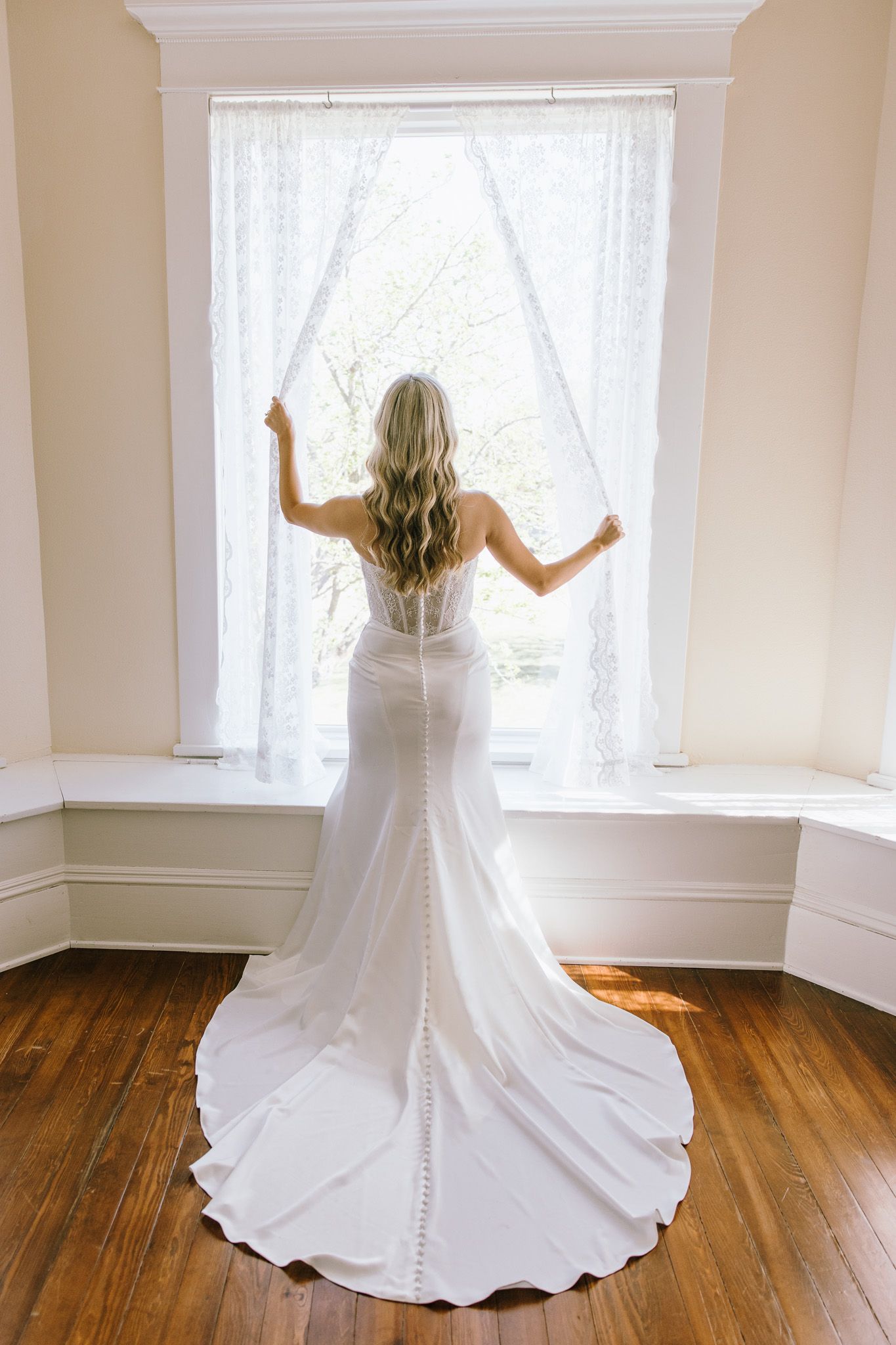 Bride in white fitted wedding gown with train stands facing window, long blonde hair cascading down her back.