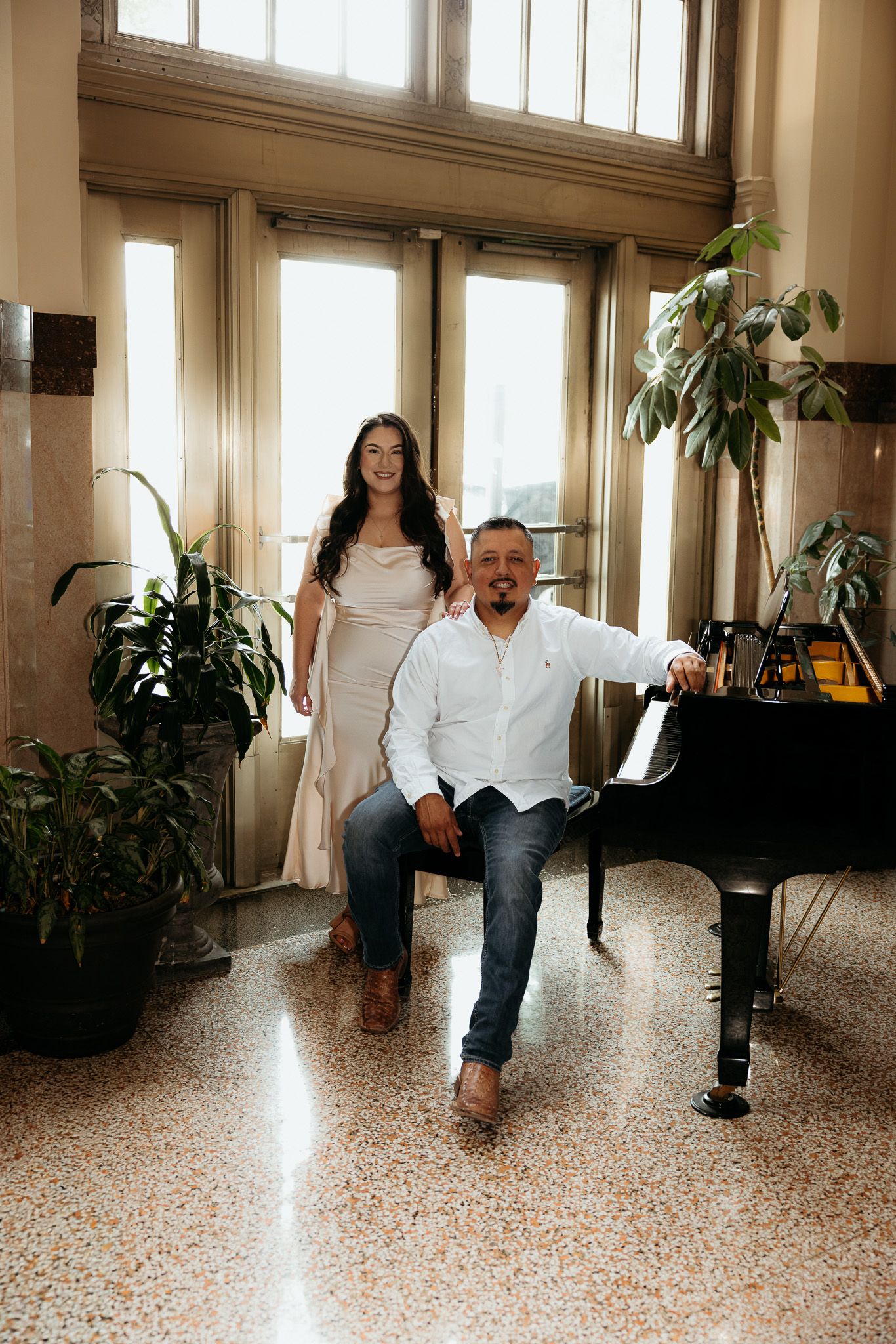 Couple posing by a grand piano in an elegant sunlit room with large windows and lush green plants.