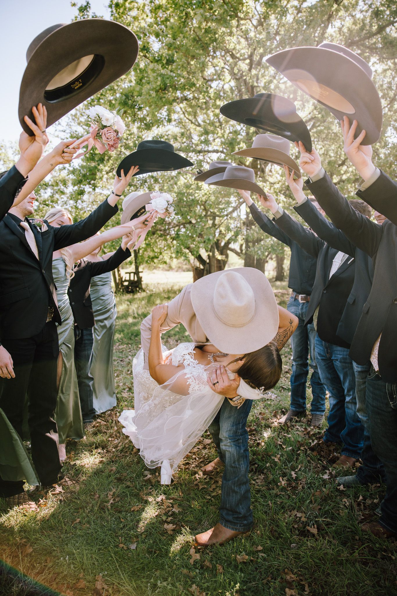 Bride and groom share a dip kiss under an arch of raised cowboy hats held by wedding party outdoors.