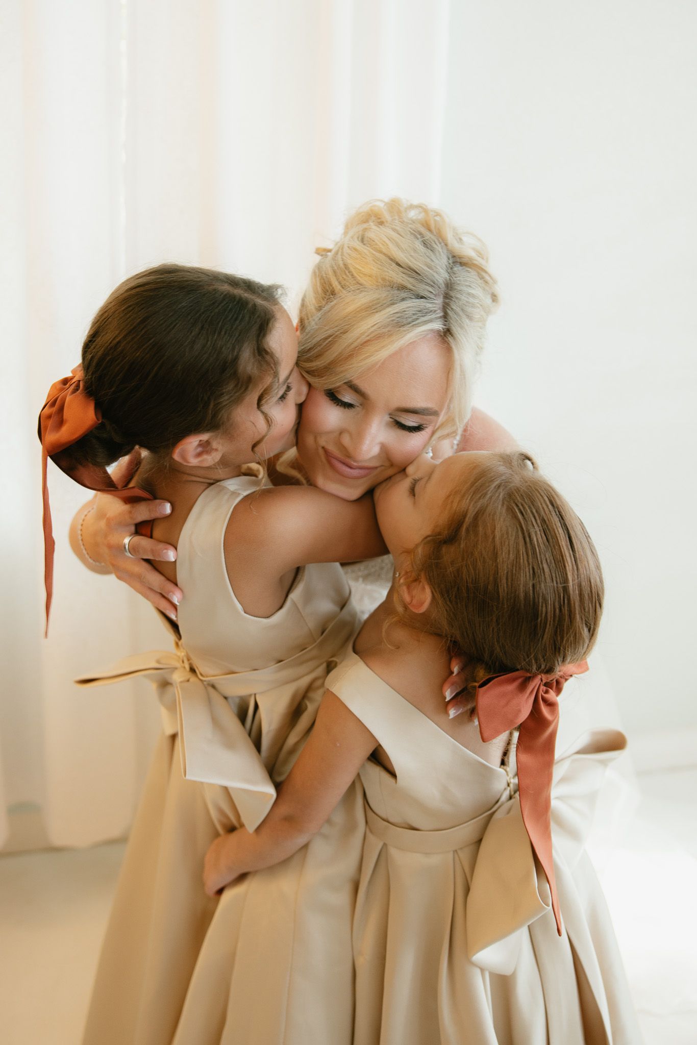 Blonde bride in white dress hugged and kissed by two flower girls in champagne dresses with pink and orange bows.