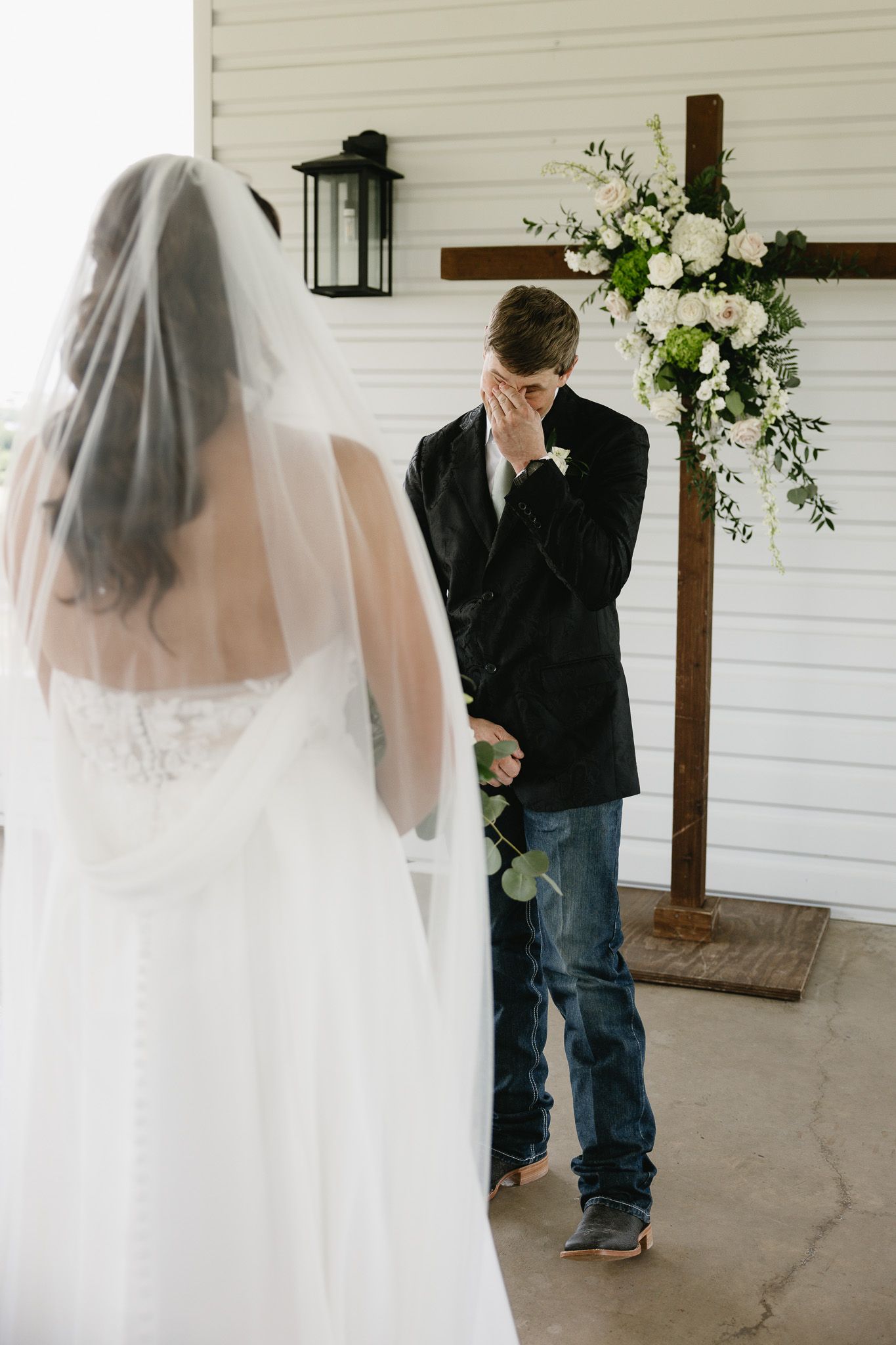 Emotional groom covering his face while seeing bride in white wedding dress and veil for first time at outdoor ceremony.