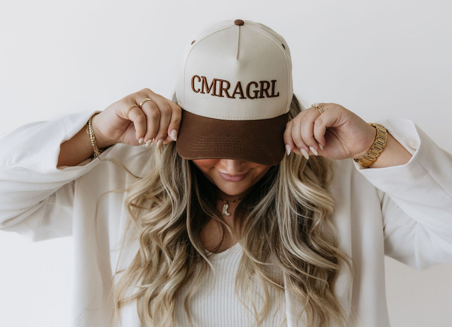 Stylish woman in baseball cap and white sweater poses against white wall.