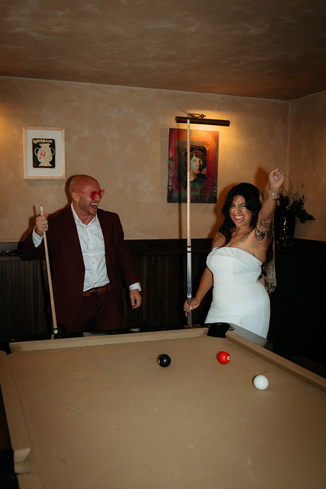 Couple celebrating while playing pool billiards, woman in white dress raising arm in victory, man in burgundy blazer holding cue stick.