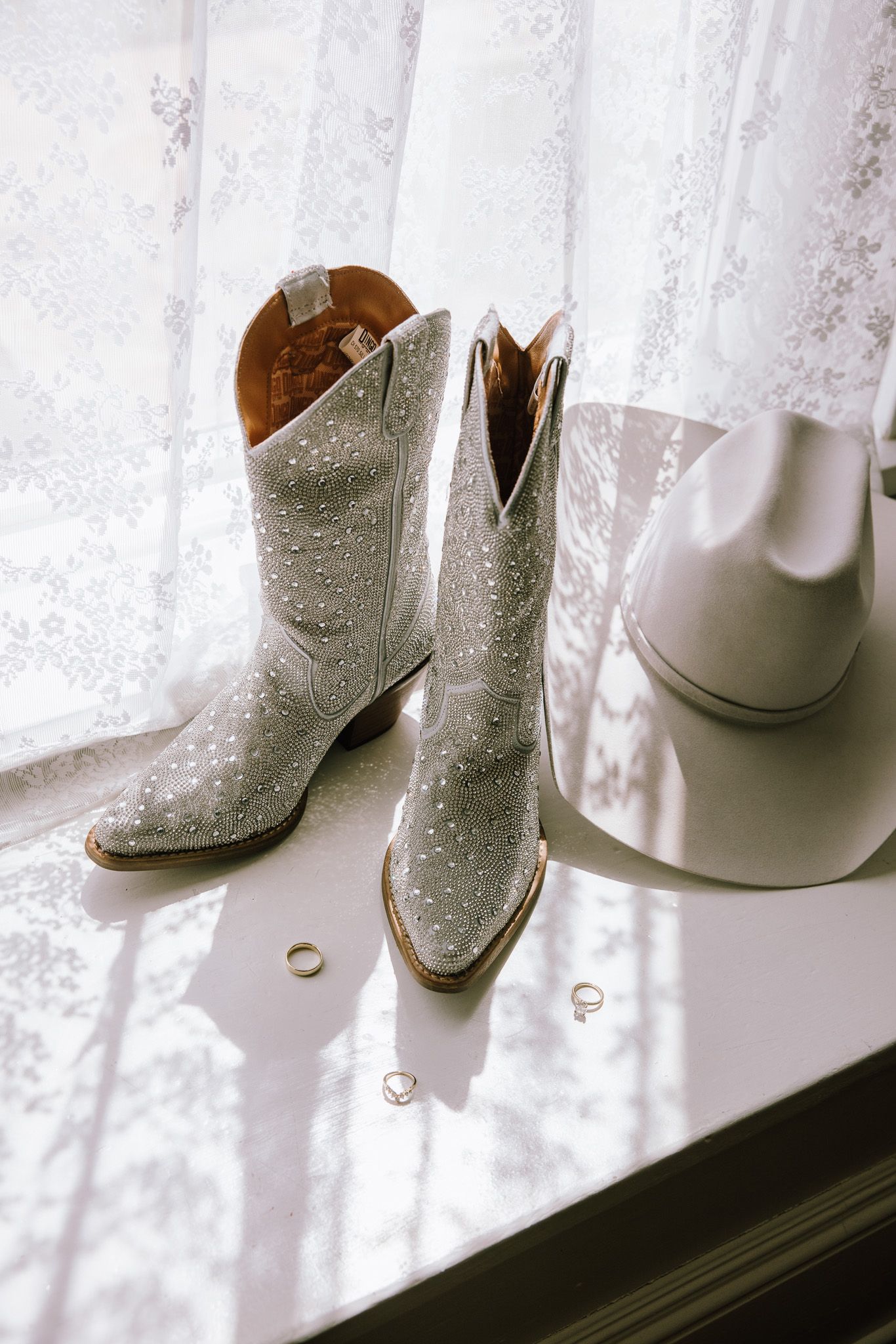 White studded cowboy boots and hat with gold rings on a windowsill with lace curtains, perfect for a country wedding.