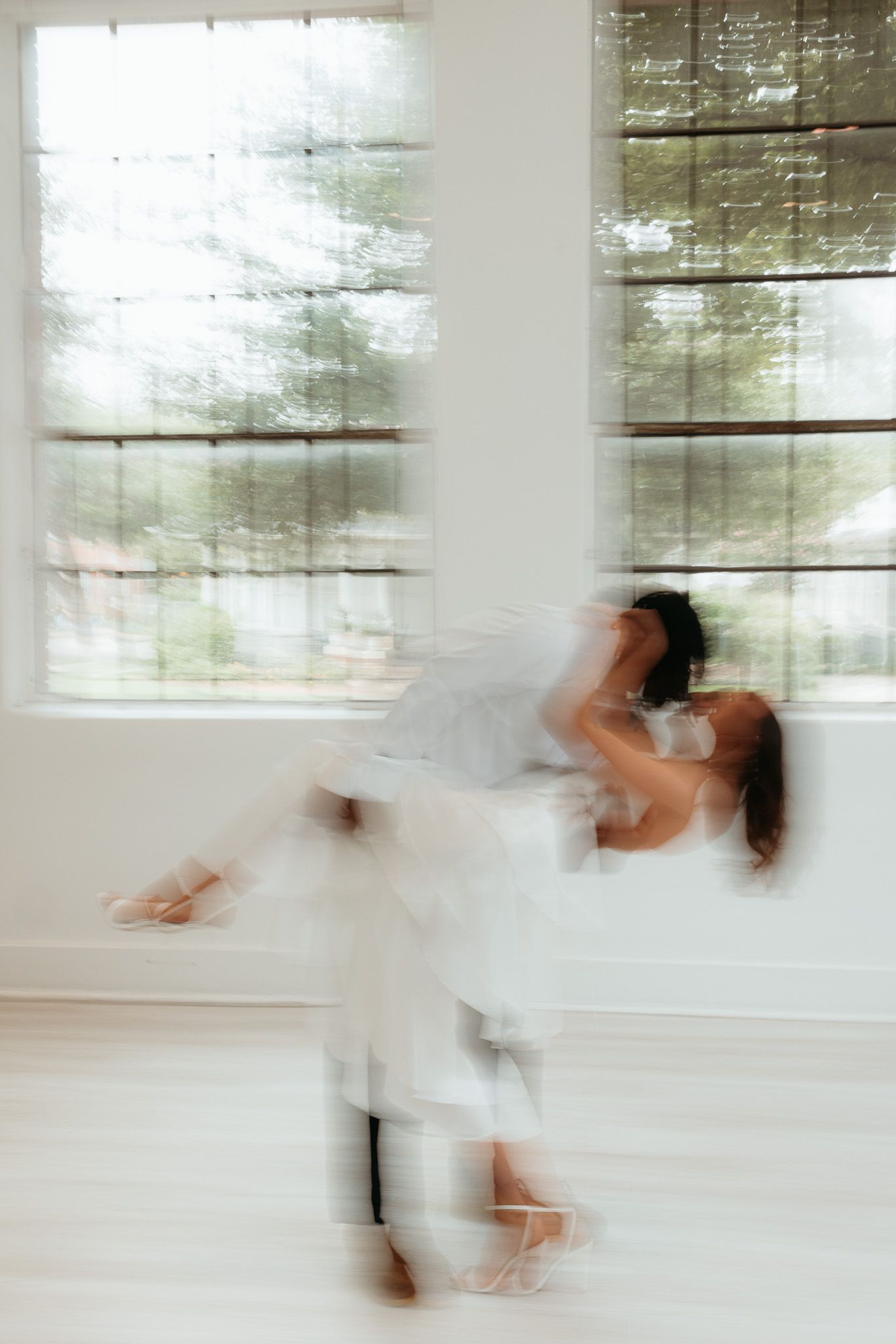 Motion blur captures dancers in ethereal white performing a graceful lift in a bright sunlit studio.