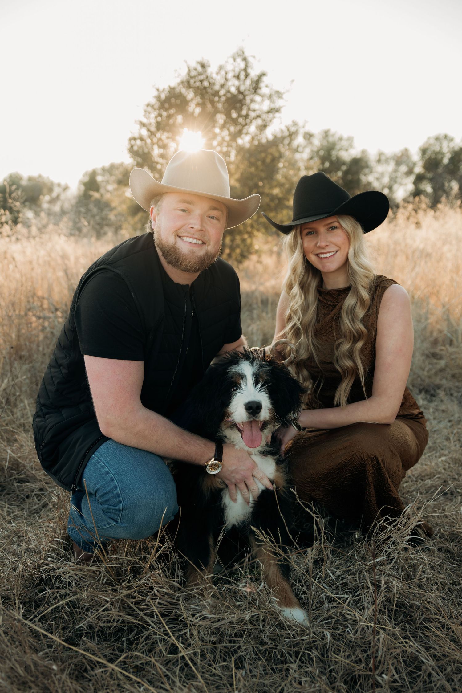 Couple in cowboy hats crouching with black and white dog in golden field at sunset.