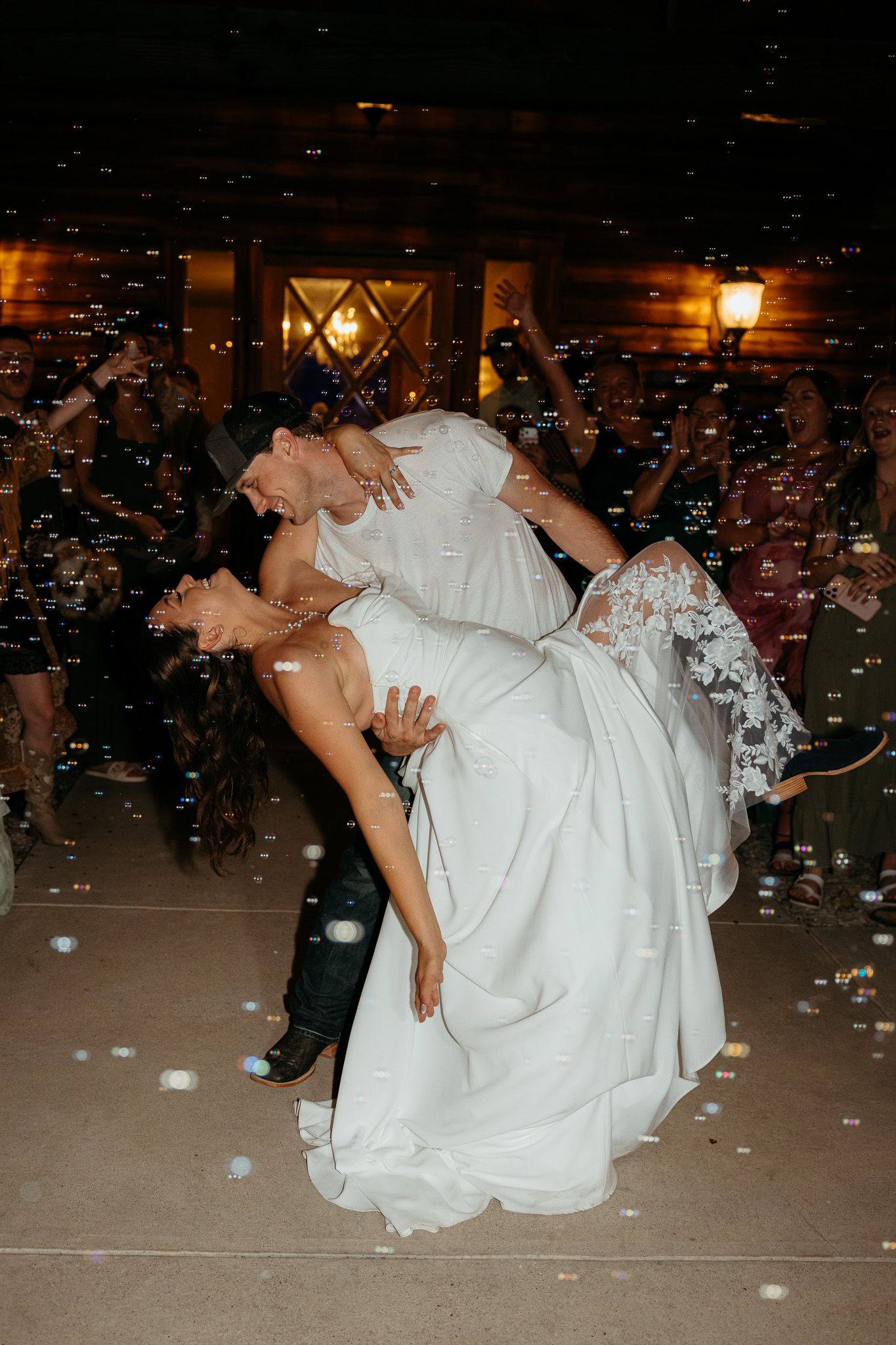 Groom dips bride in white lace wedding dress on dance floor surrounded by guests and floating bubbles at night.