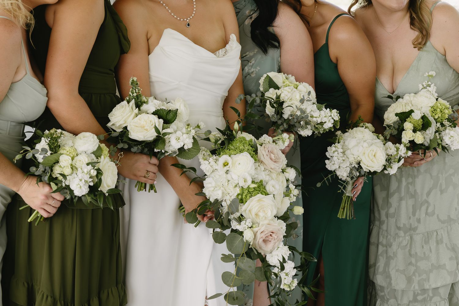 Bride in white gown with bridesmaids in green dresses holding white and blush floral bouquets with eucalyptus greenery.