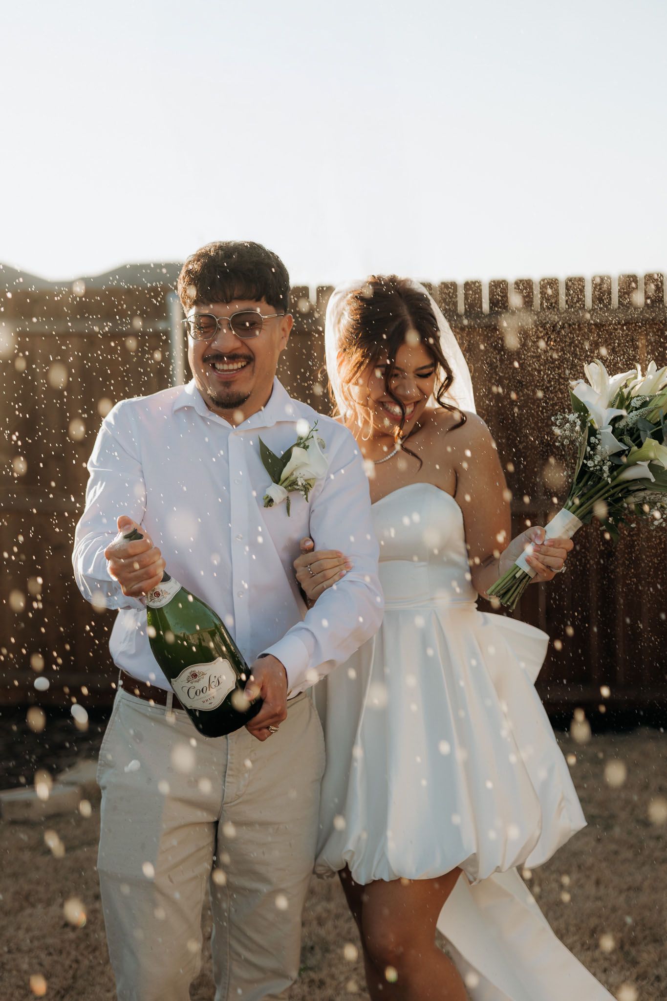 Newlywed couple celebrating with champagne spray, bride in white mini dress holding flowers, groom in light blue suit.