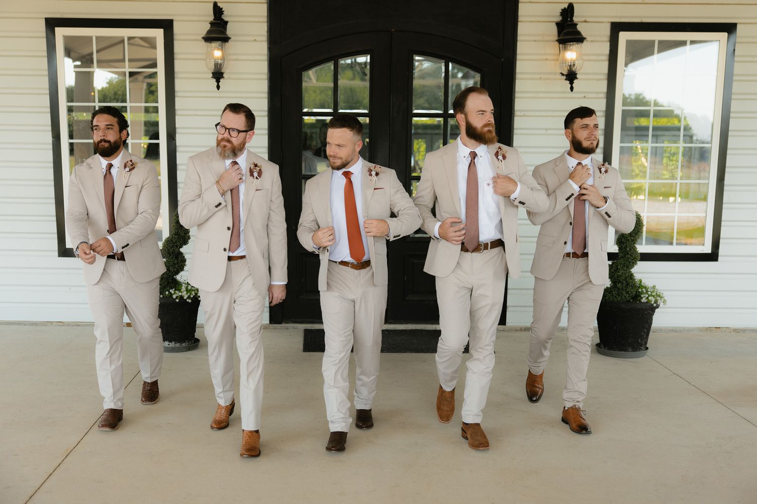 Groom and groomsmen in tan suits with orange ties walking together at modern wedding venue with black framed windows.