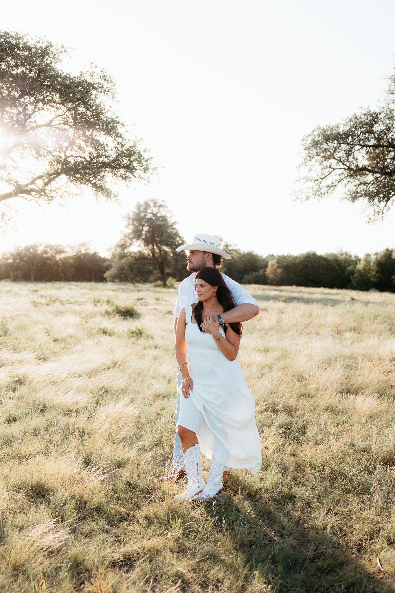 Couple embracing in a sunlit field, woman in white dress and boots, man in white hat, golden hour outdoor portrait.