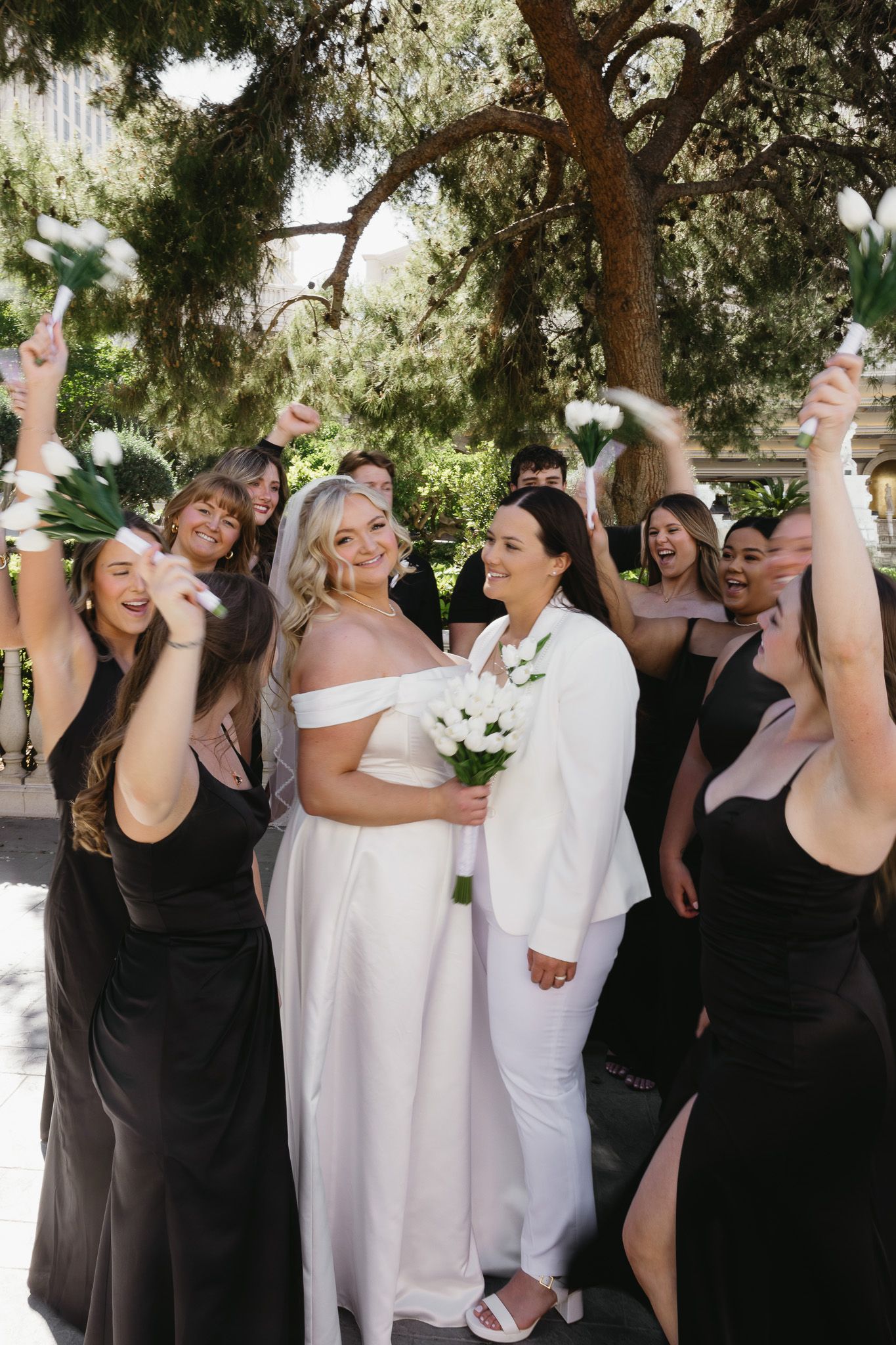 Two brides in white celebrate with bridesmaids in black dresses, holding bouquets outdoors under trees.