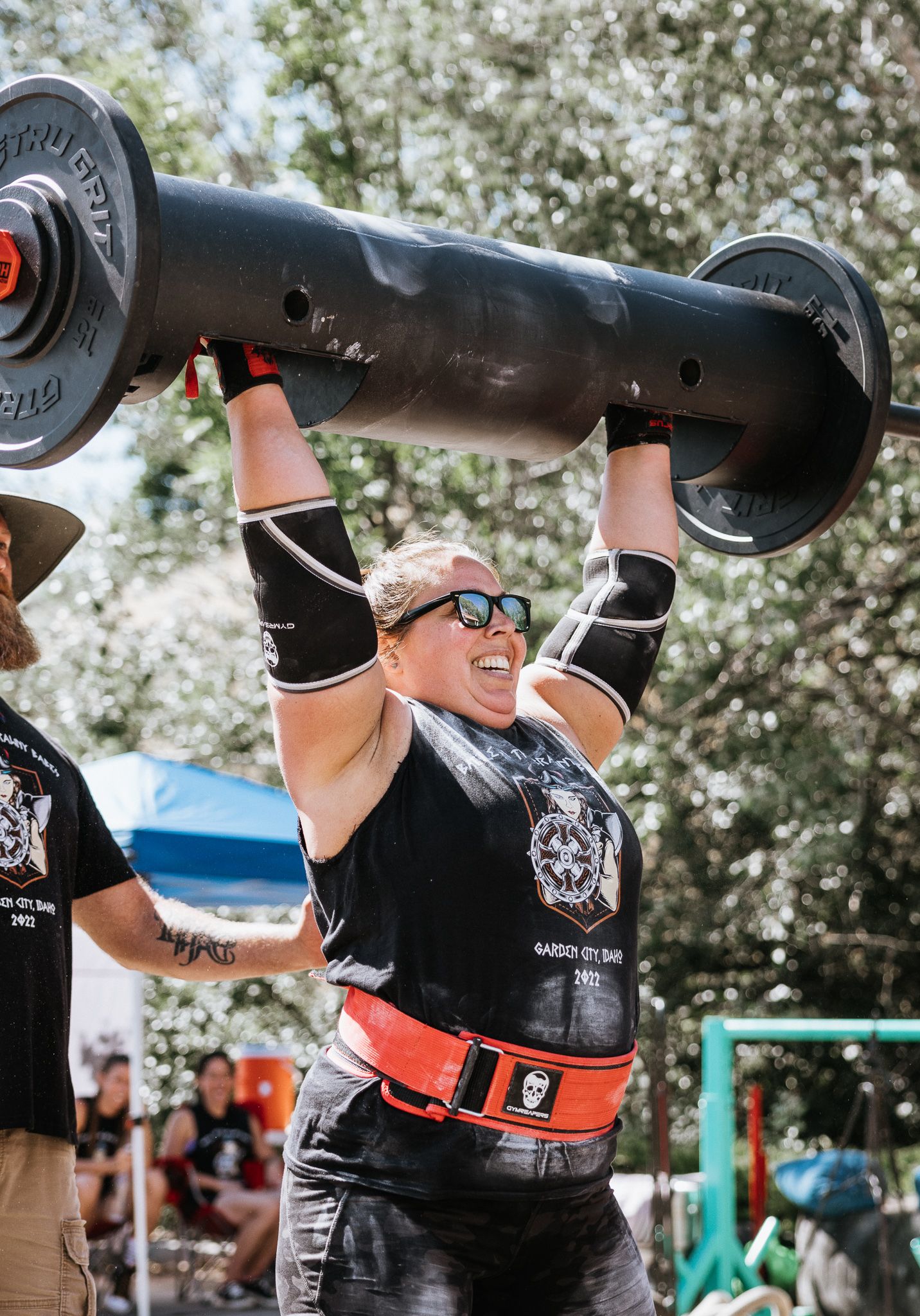 Competitors lifting heavy black barrels overhead during an outdoor strength competition.