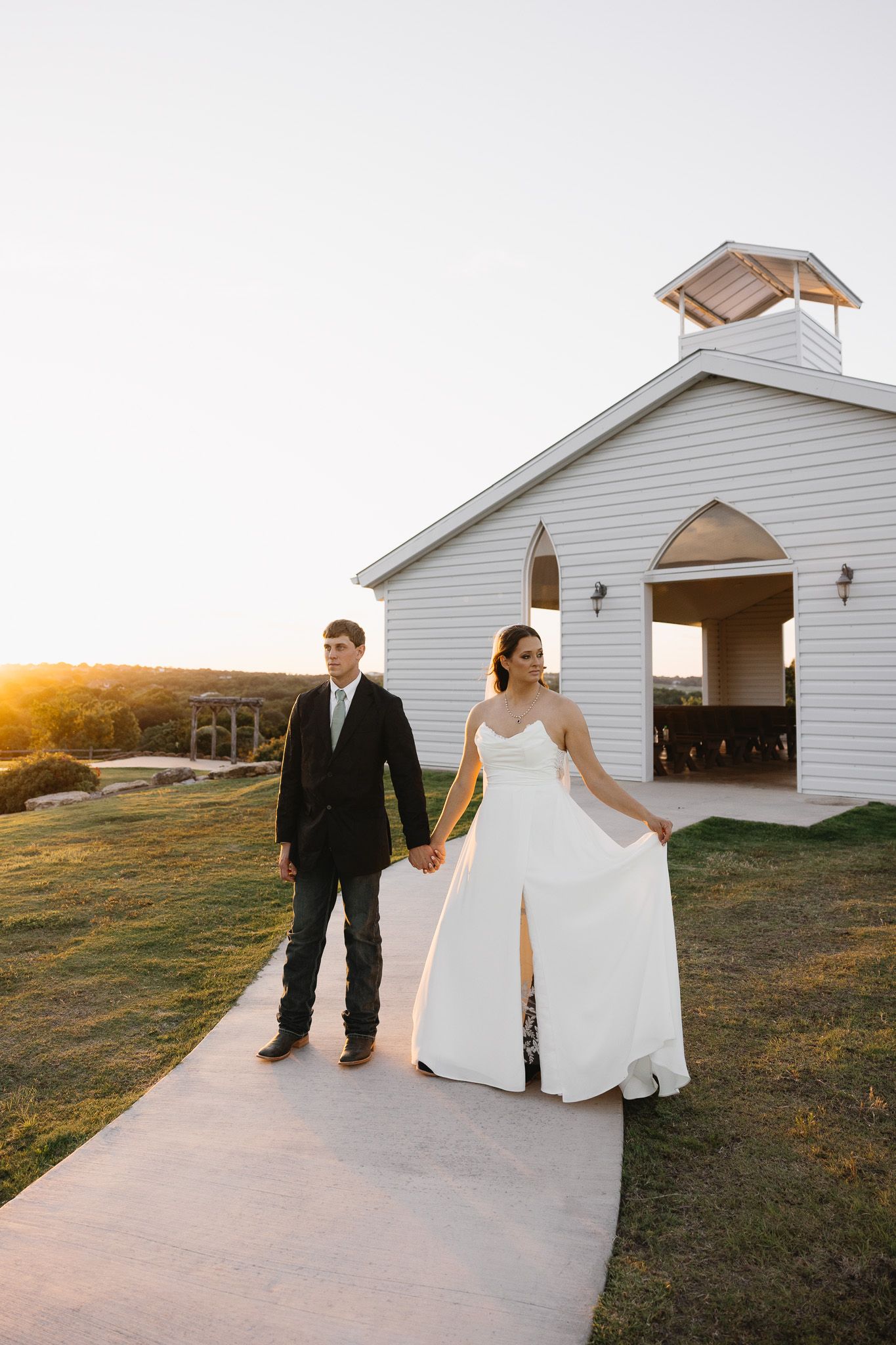 Bride and groom hold hands walking from a white chapel at golden hour on their wedding day.