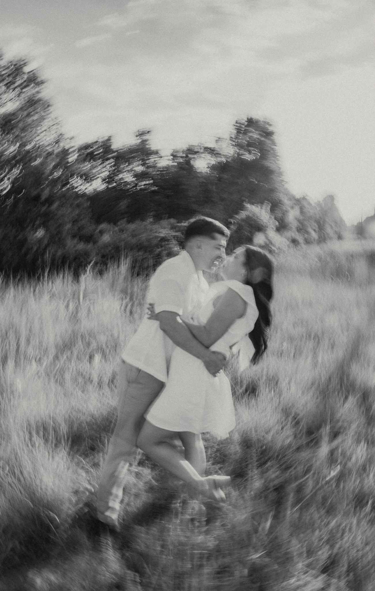 A romantic couple embraces and dances in a field of tall grass at sunset in black and white.