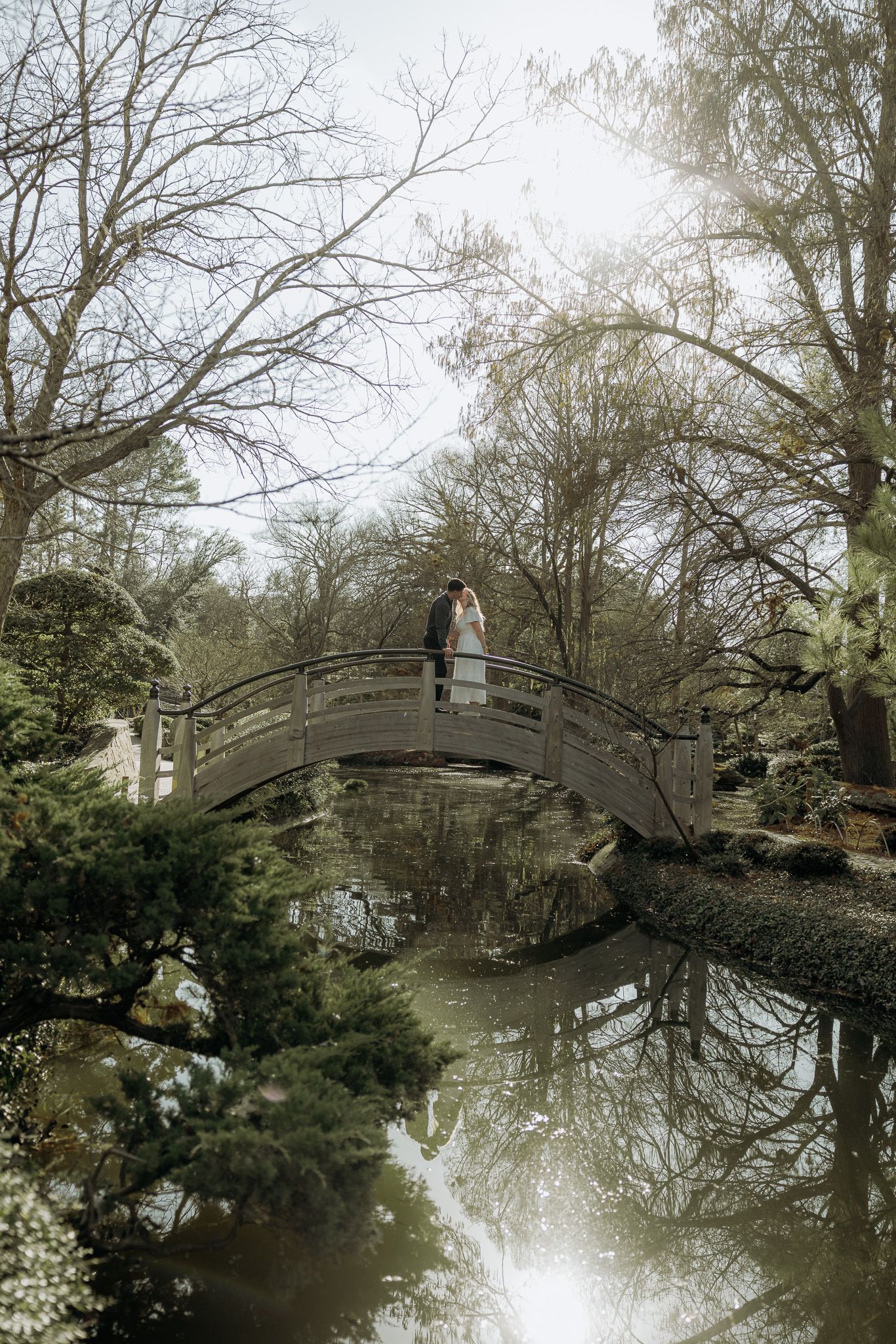 A curved wooden footbridge spans across a tranquil pond surrounded by bare winter trees reflecting in the water.